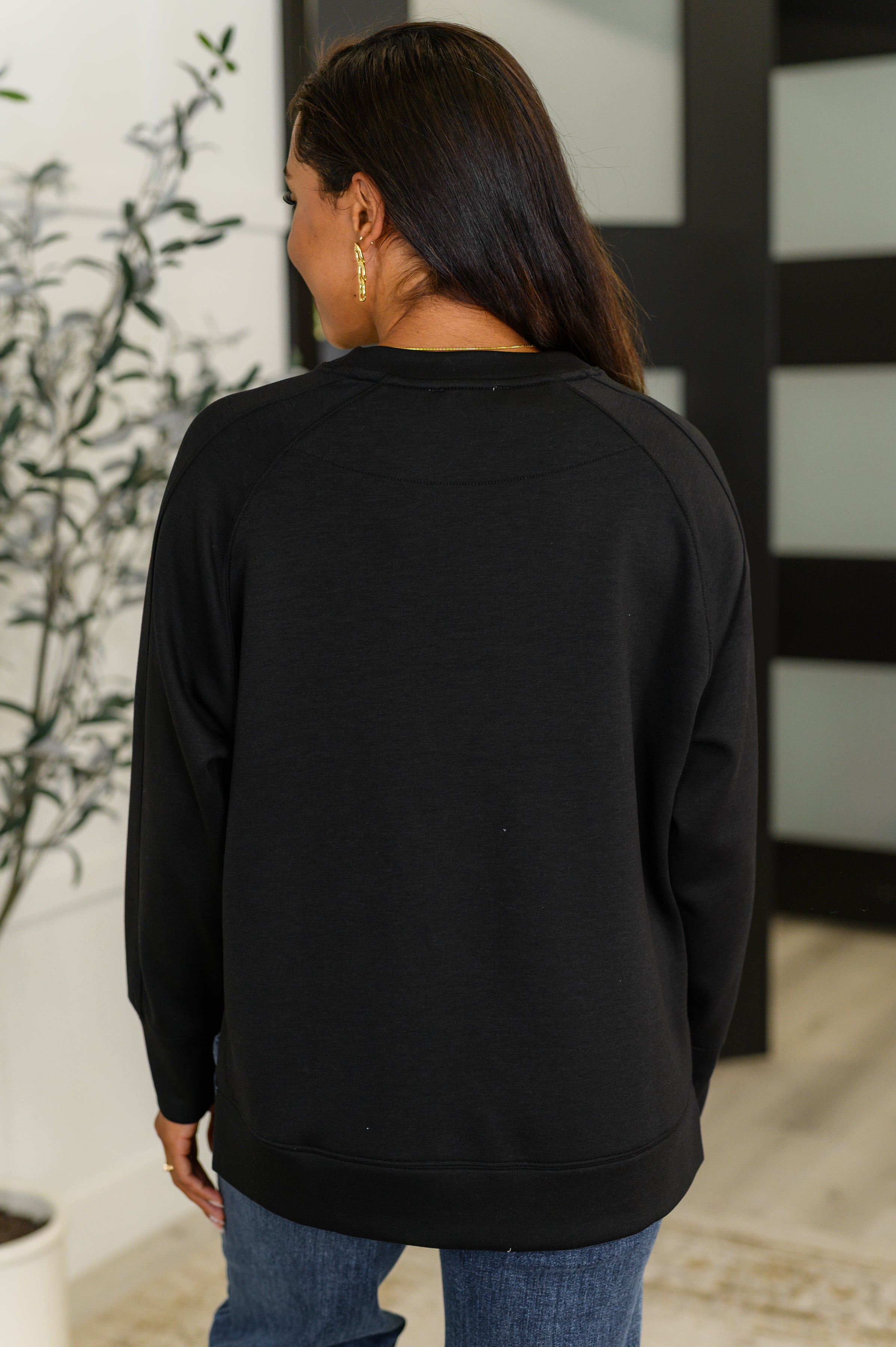Woman wearing black relaxed scuba pullover viewed from the back, paired with blue jeans indoors