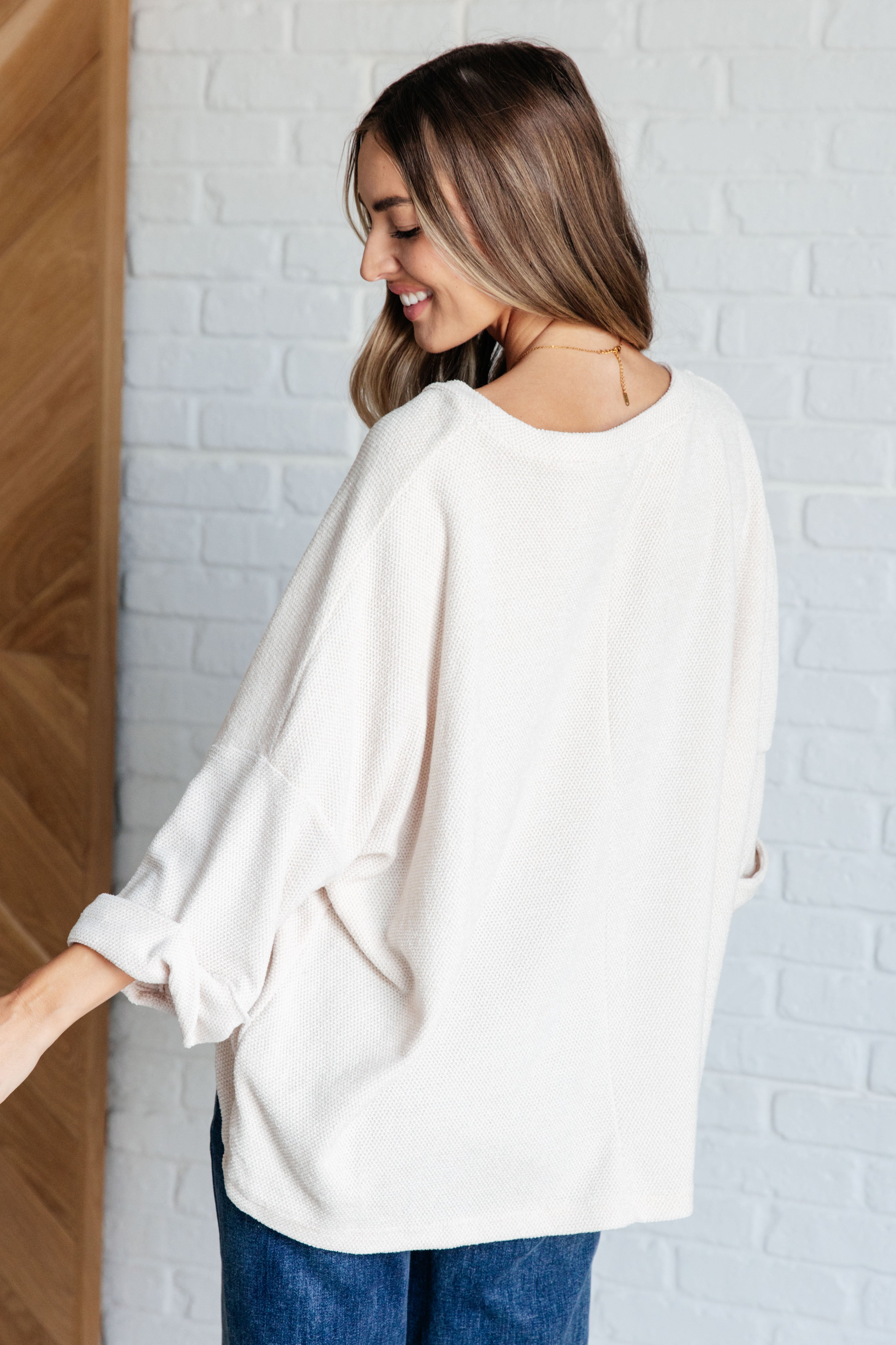 Woman smiling wearing Ivory Thoughts chenille blouse with loose fit and rolled sleeves standing against white brick wall