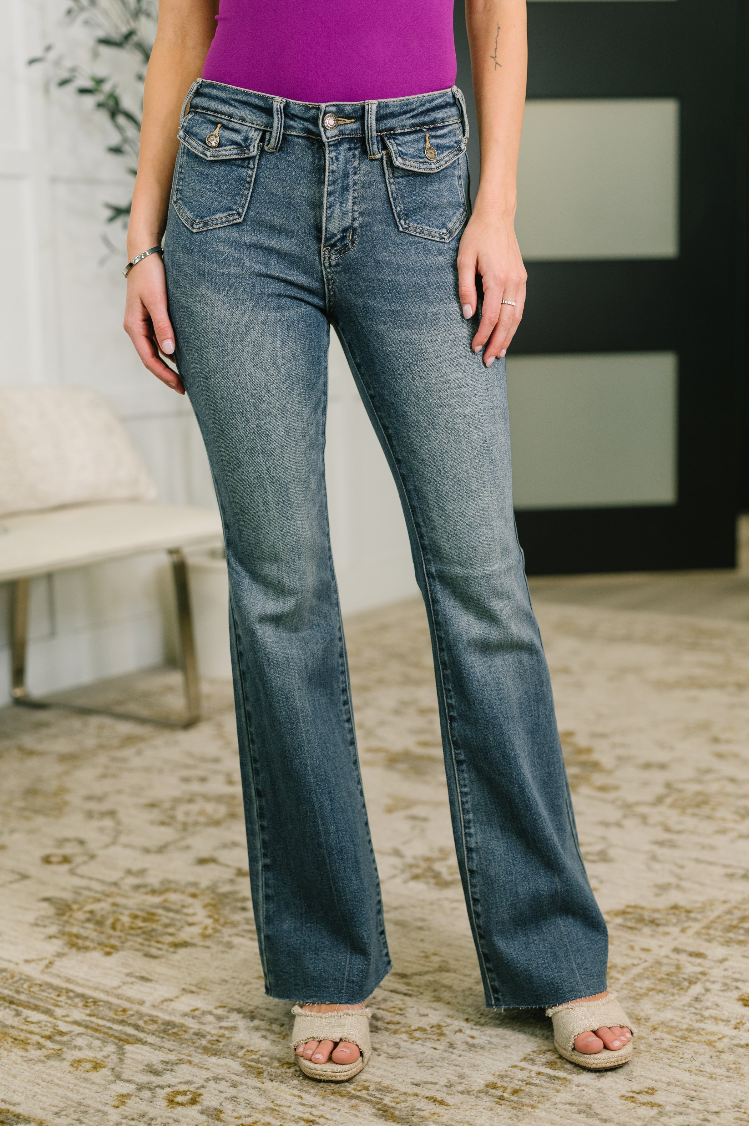 Mid rise blue flare jeans with front flap pockets worn with purple top and open-toe woven sandals indoors