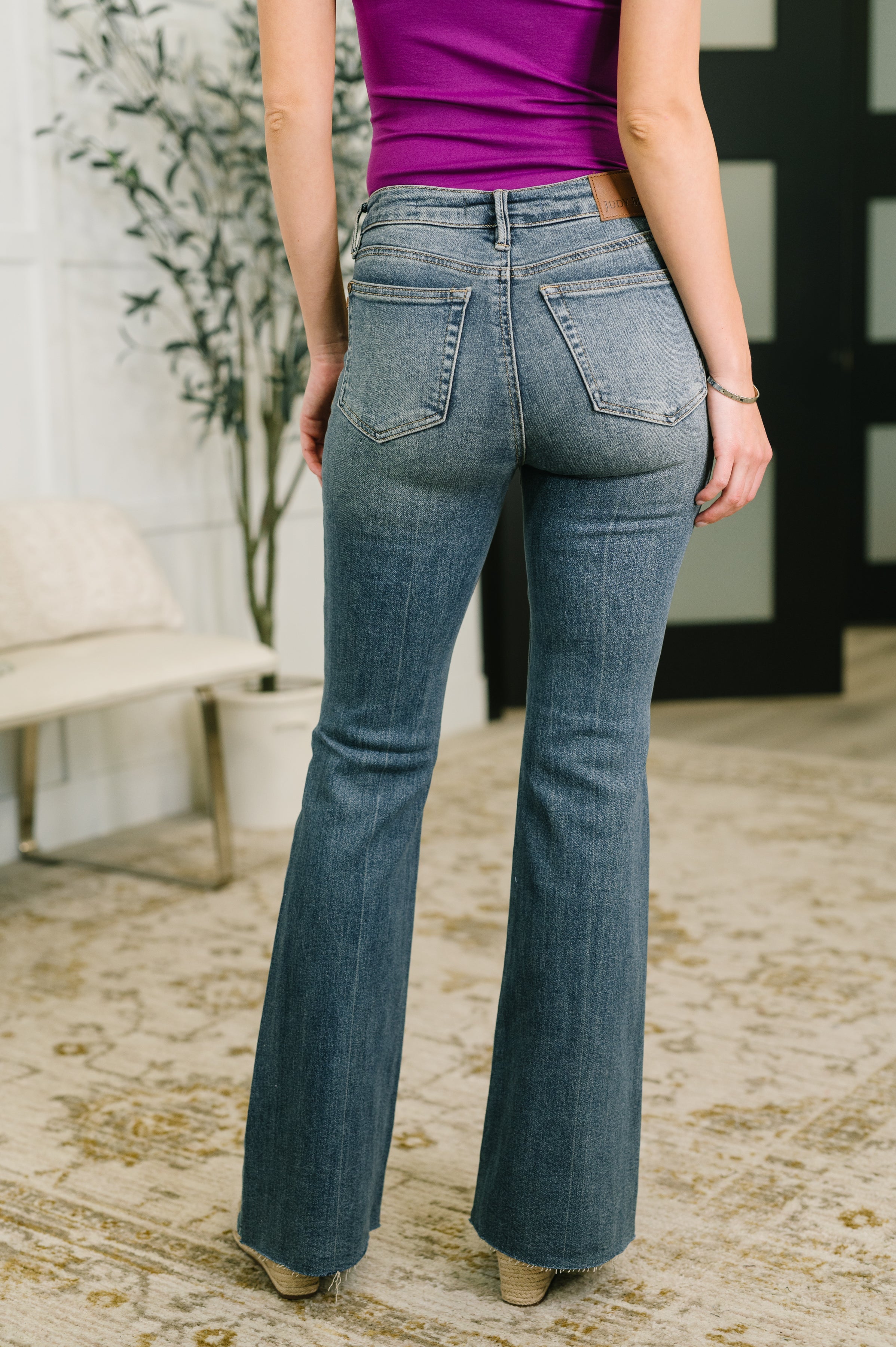 Woman wearing blue mid rise flare jeans with flap pockets paired with a purple top indoors on patterned rug