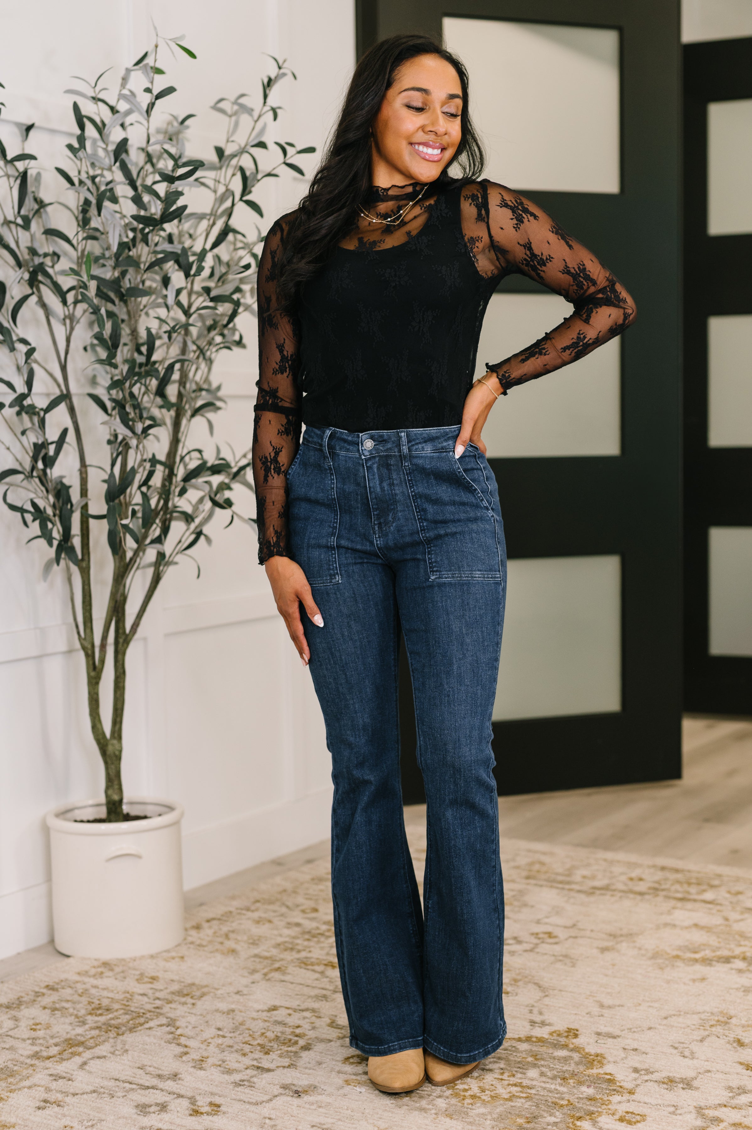 Woman standing indoors wearing blue Jada high waist flare jeans with patch pockets paired with black sheer lace top