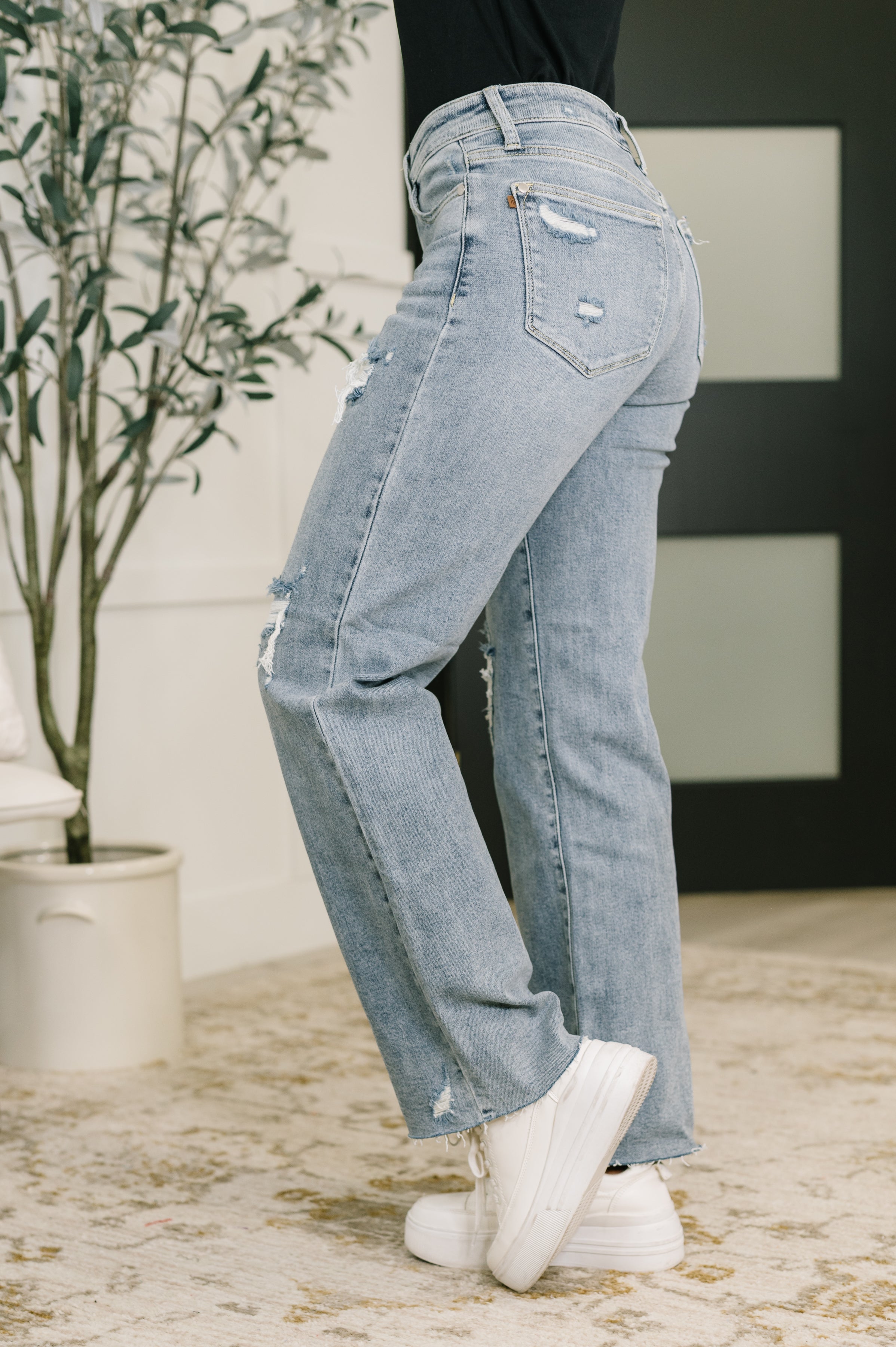 Side angle of woman wearing blue Jade Mid Rise 90's Straight jeans with distressed detailing and white sneakers indoors
