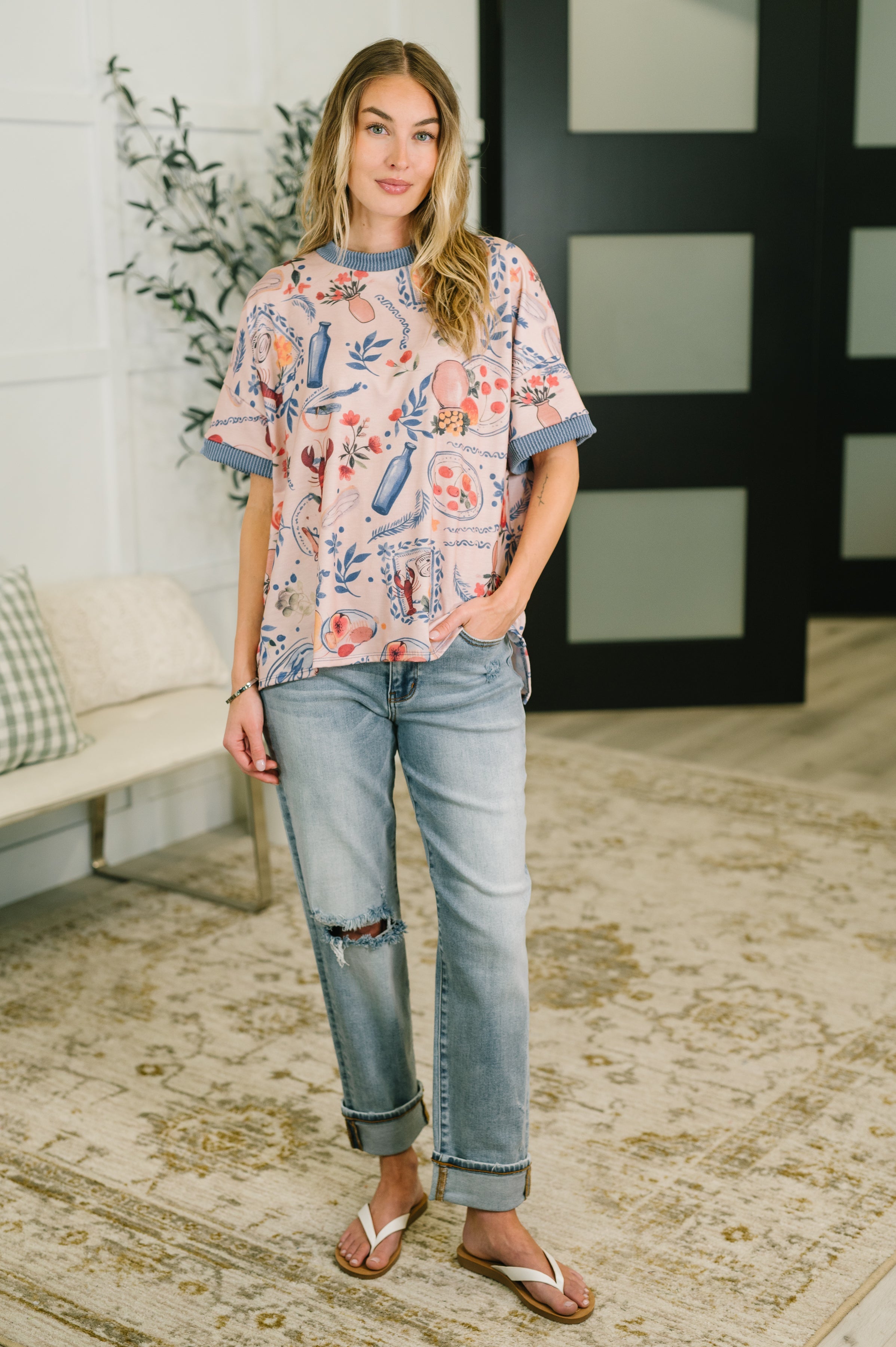 Woman wearing a loose patterned pastel top with rolled light wash jeans and white sandals standing indoors on a patterned rug