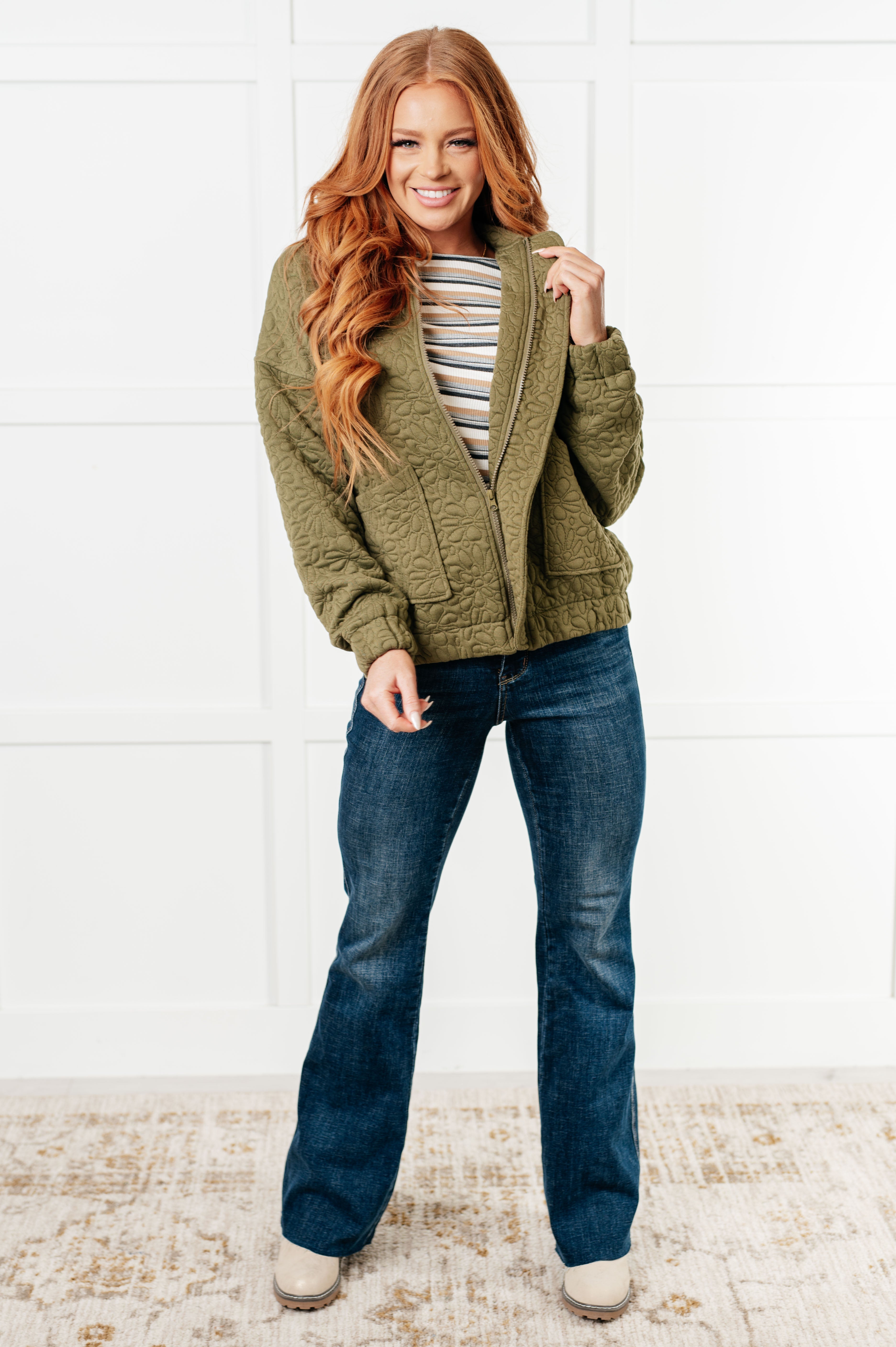Woman wearing an olive green floral quilted zip up jacket over a grey striped shirt, blue jeans and cream boots standing against a white wall.