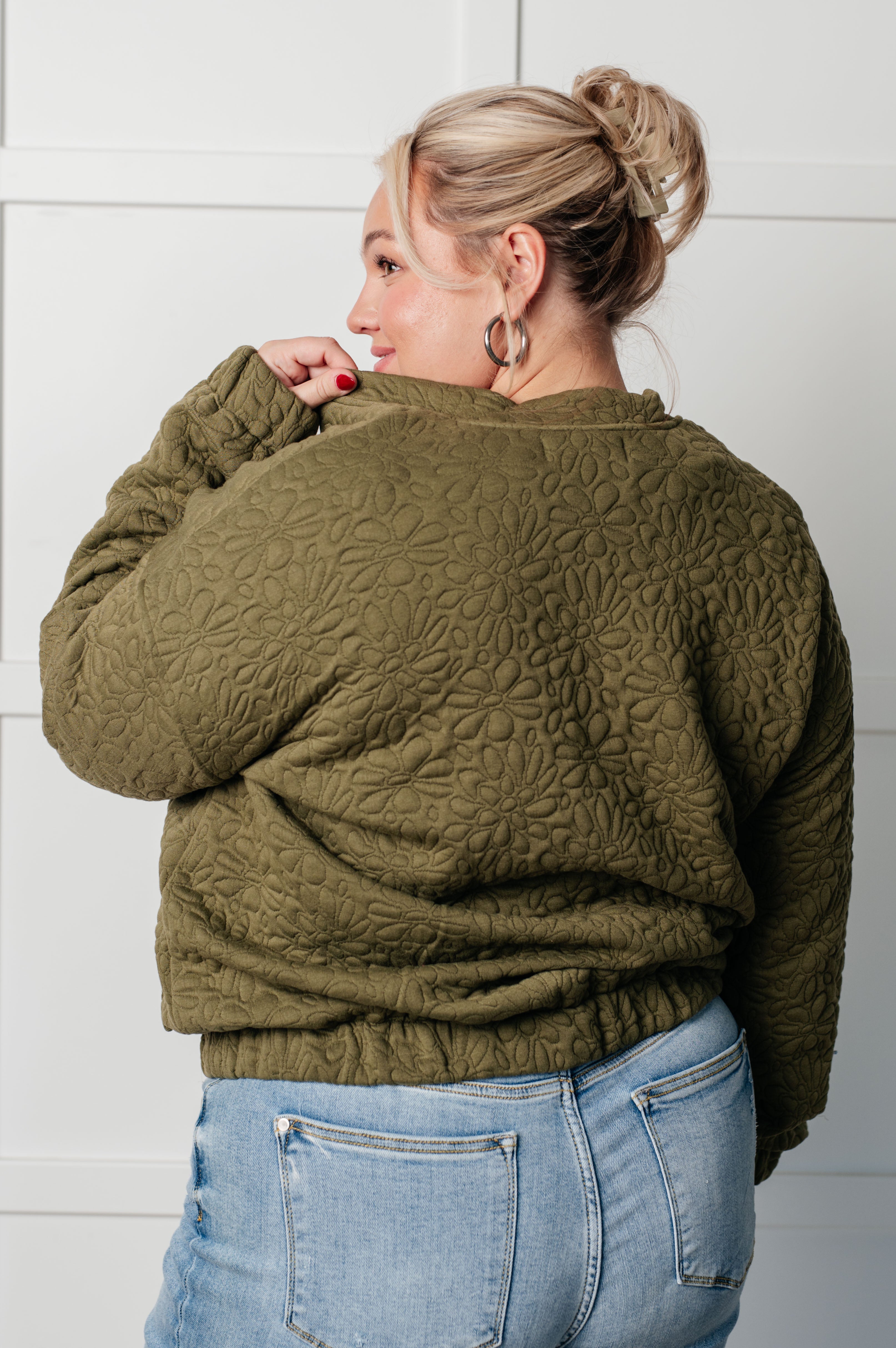 Woman wearing an olive green floral quilted zip up jacket and blue jeans standing against a white wall.
