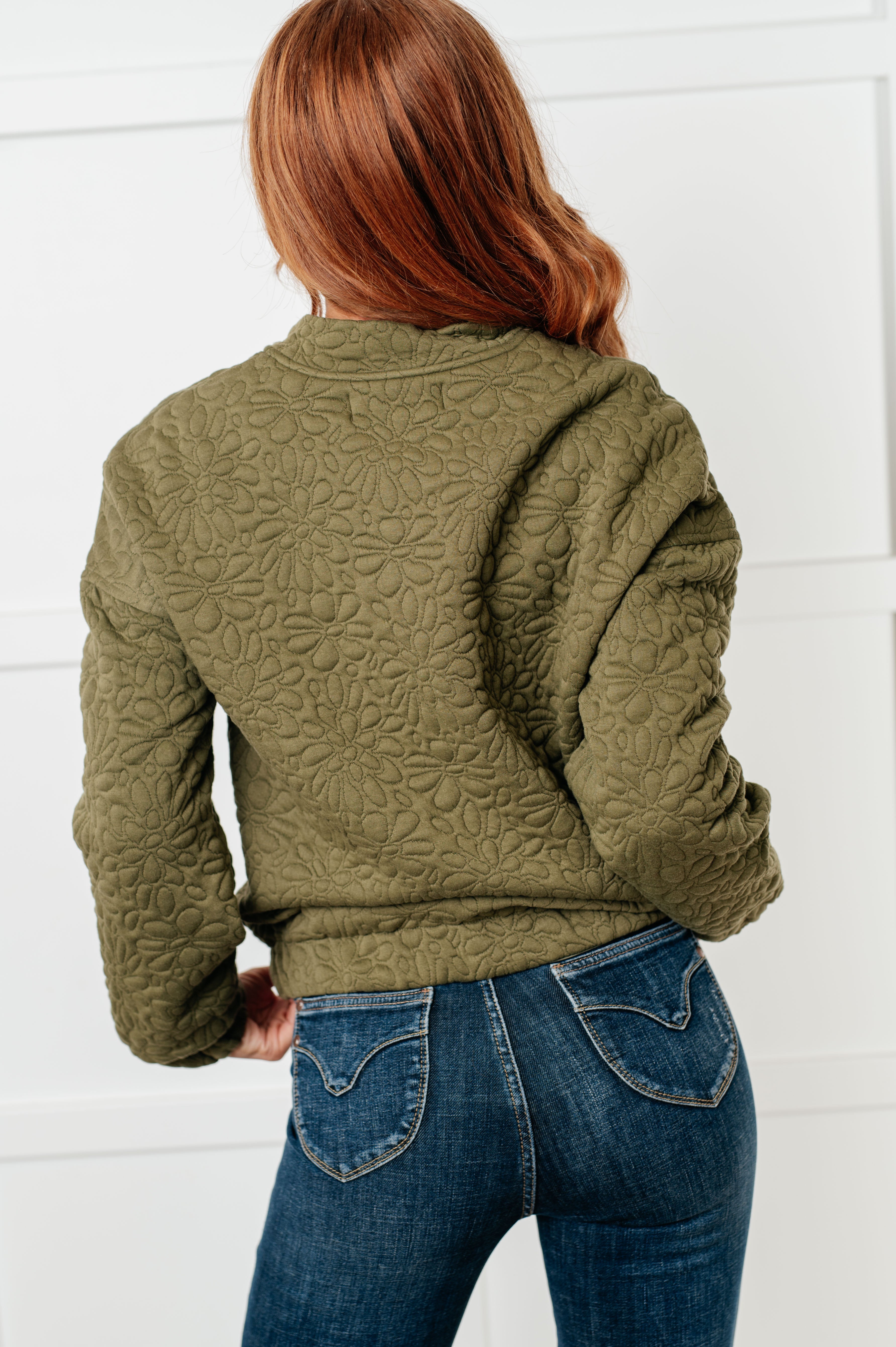 Woman wearing an olive green floral quilted zip up jacket and blue jeans standing against a white wall.