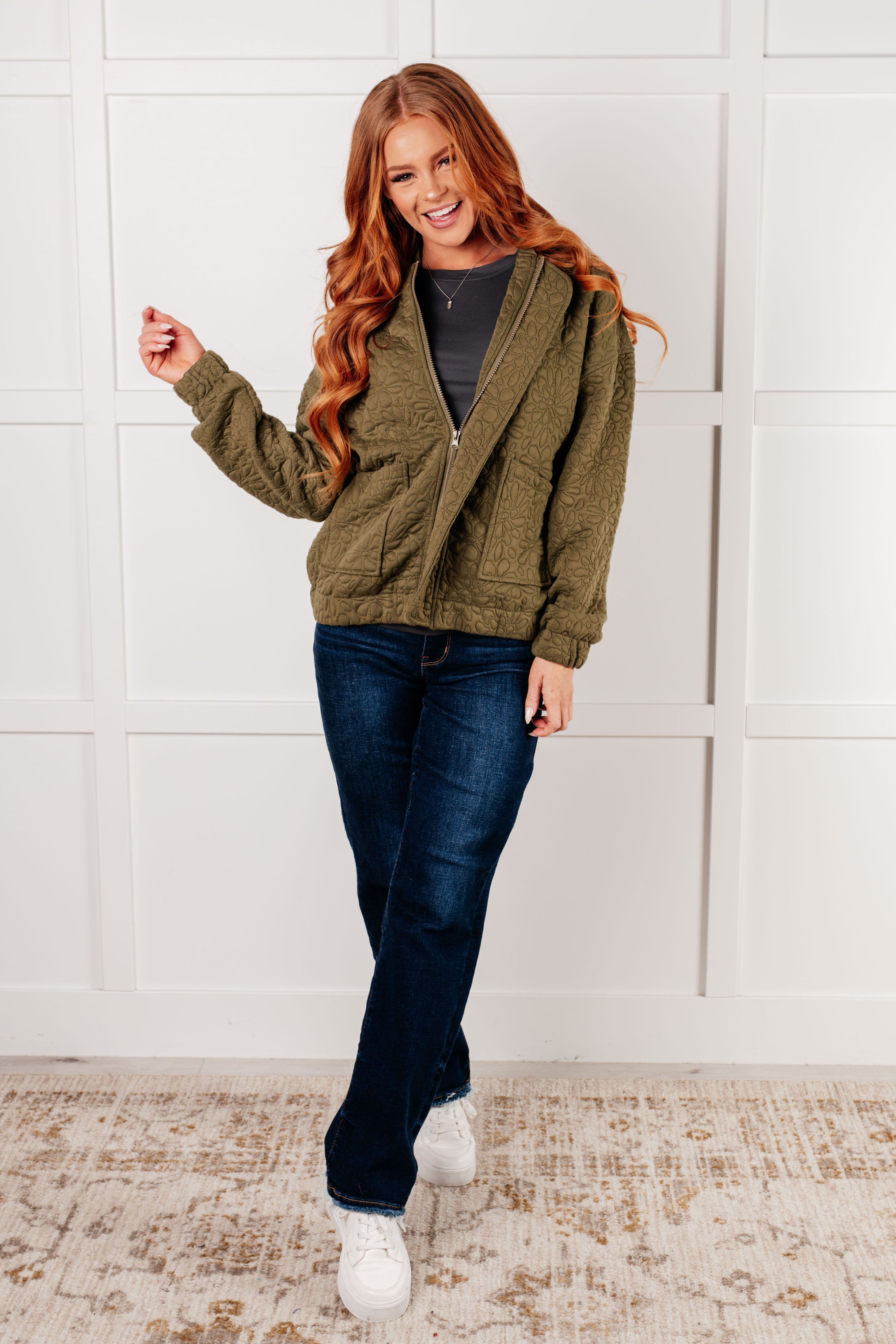 Woman wearing an olive green floral quilted zip up jacket over a charcoal shirt, blue jeans and white sneakers standing against a white wall.