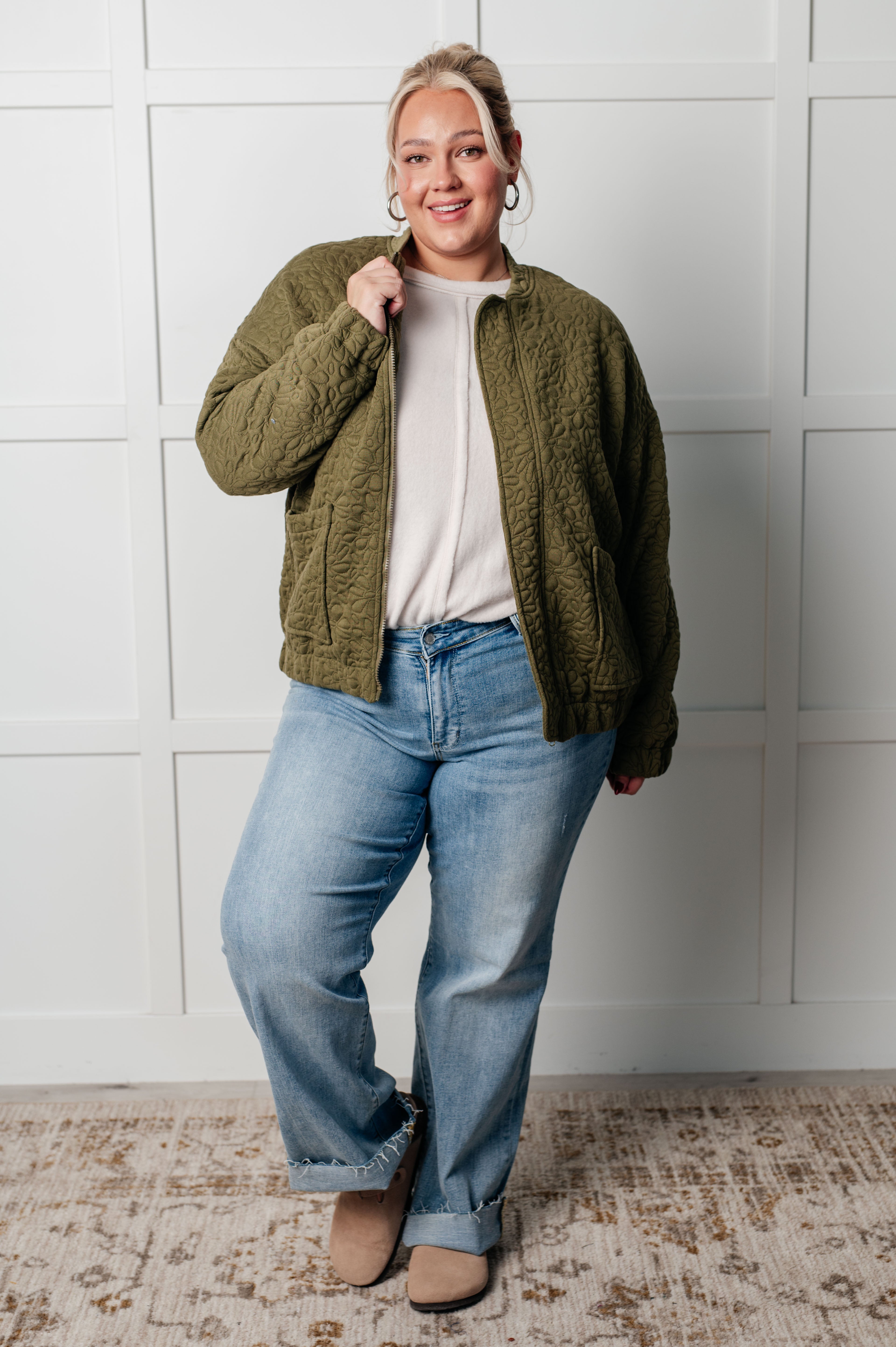 Woman wearing an olive green floral quilted zip up jacket over a white shirt, blue jeans and tan boots standing against a white wall.