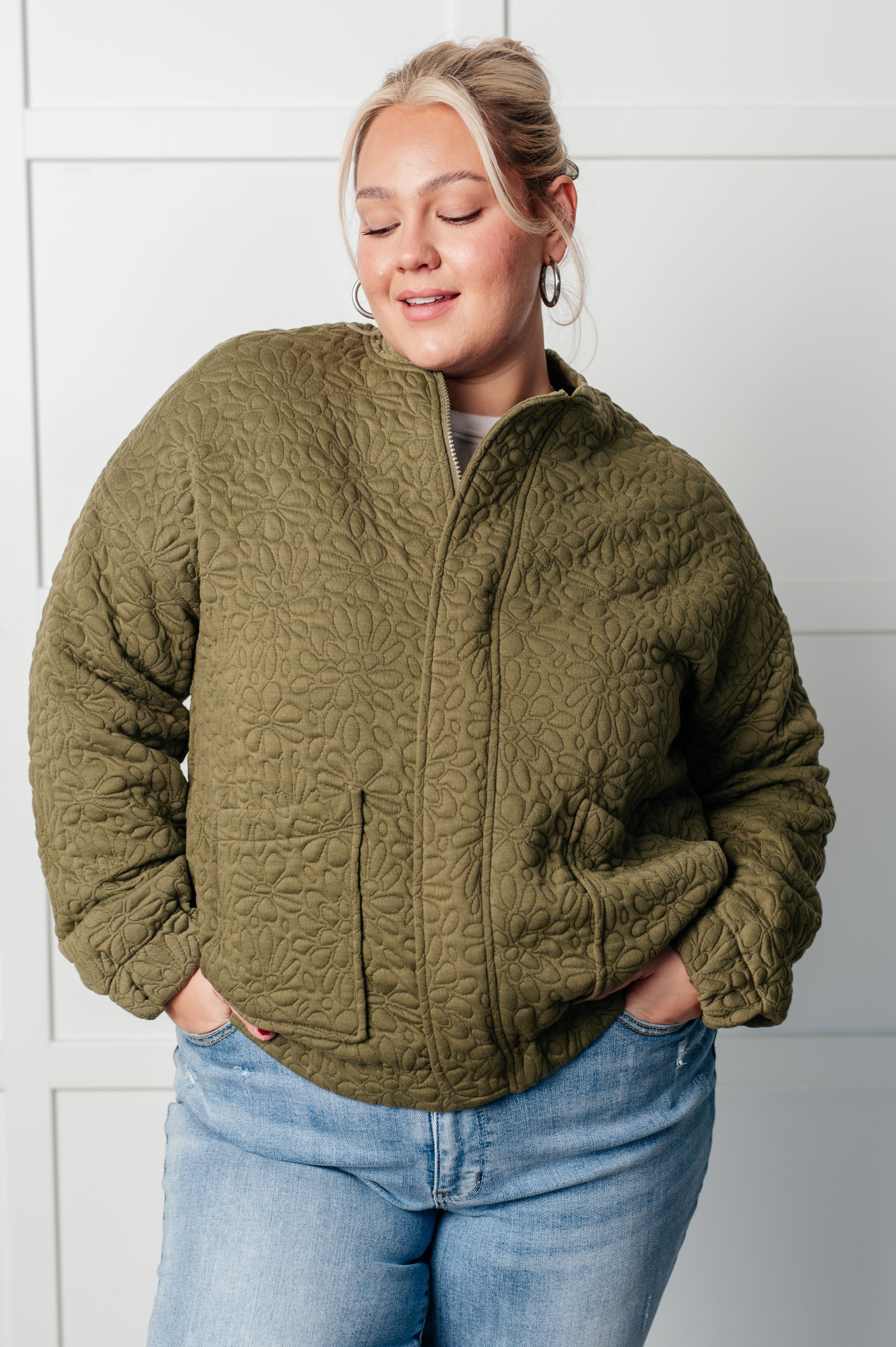 Woman wearing an olive green floral quilted zip up jacket and blue jeans standing against a white wall.