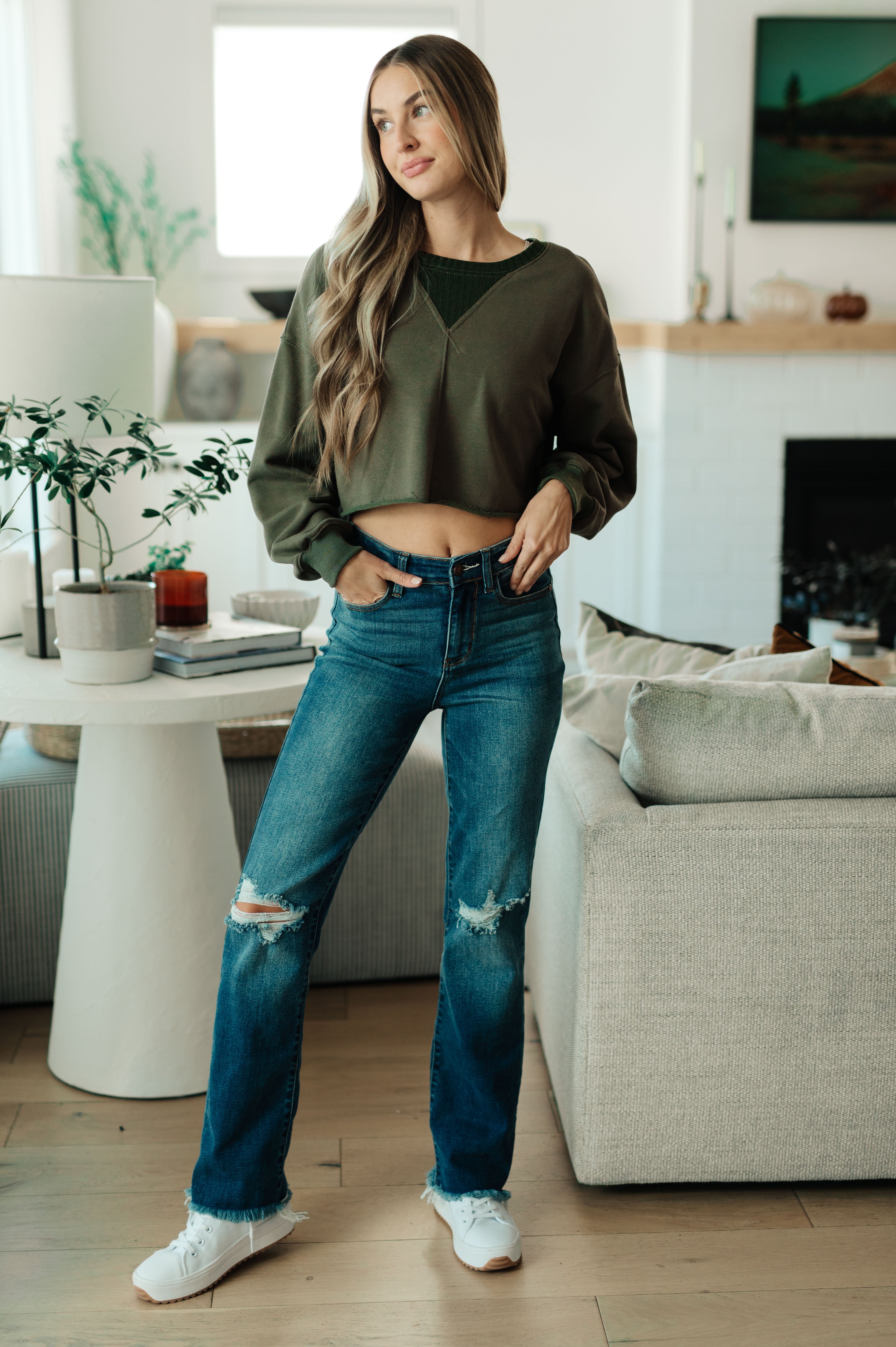 Woman indoors wearing green Just a Casual Crop Pullover layered over black top paired with ripped straight-leg blue jeans and white sneakers