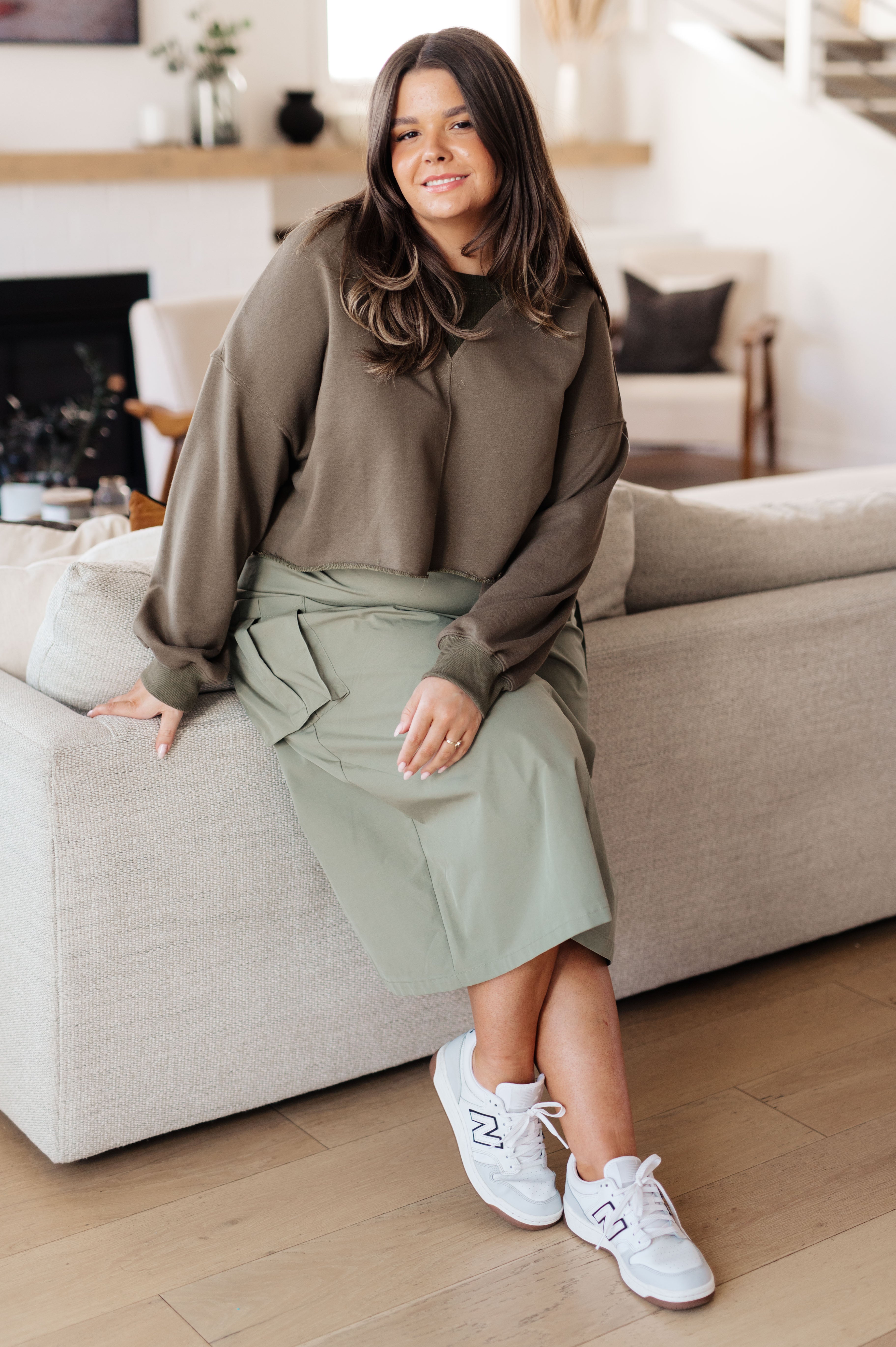 Woman seated indoors wearing green Just a Casual Crop Pullover with relaxed sleeves and light green midi skirt paired with white sneakers
