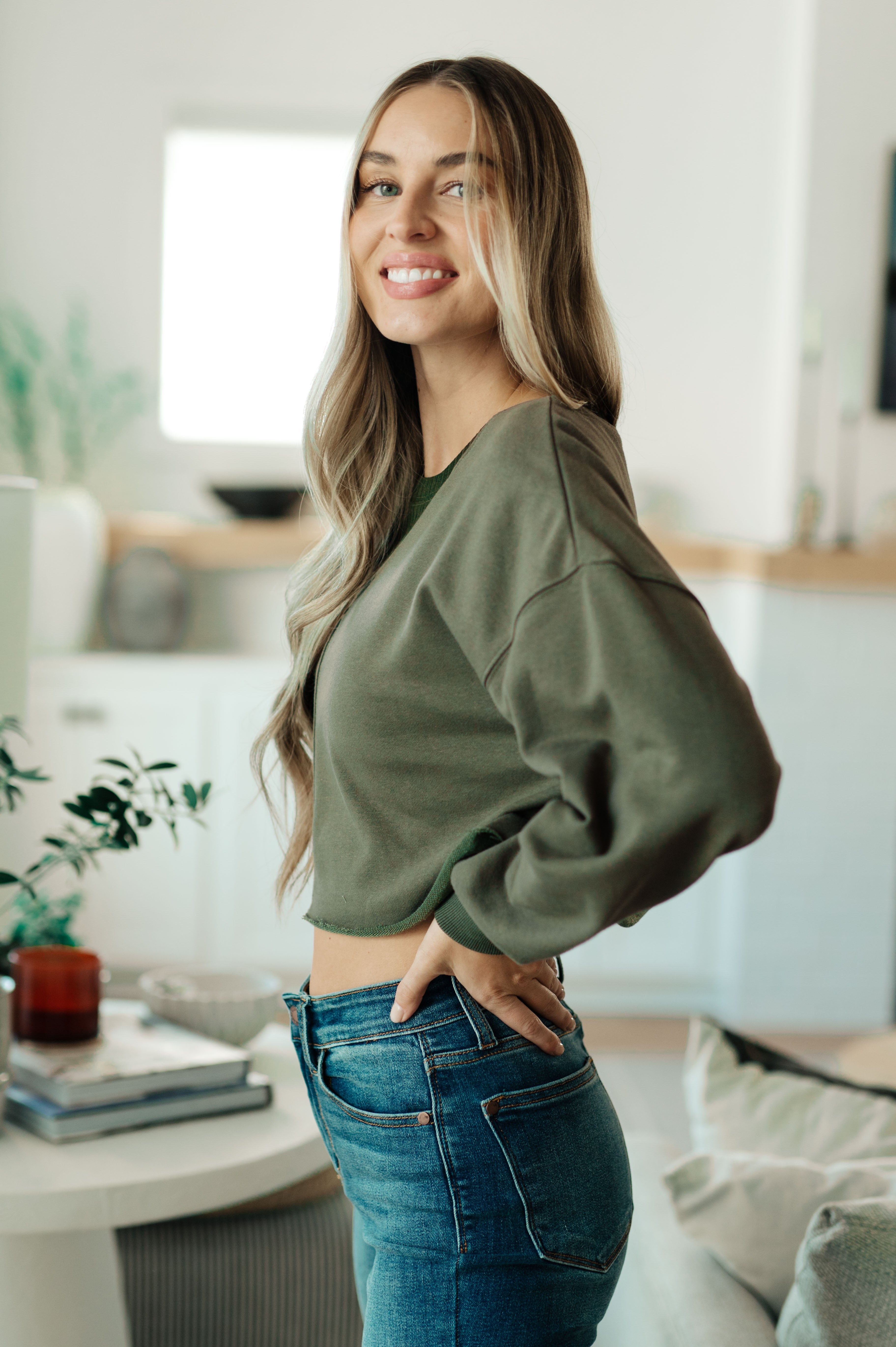 Woman indoors smiling wearing green Just a Casual Crop Pullover with loose sleeves and high-waisted blue jeans
