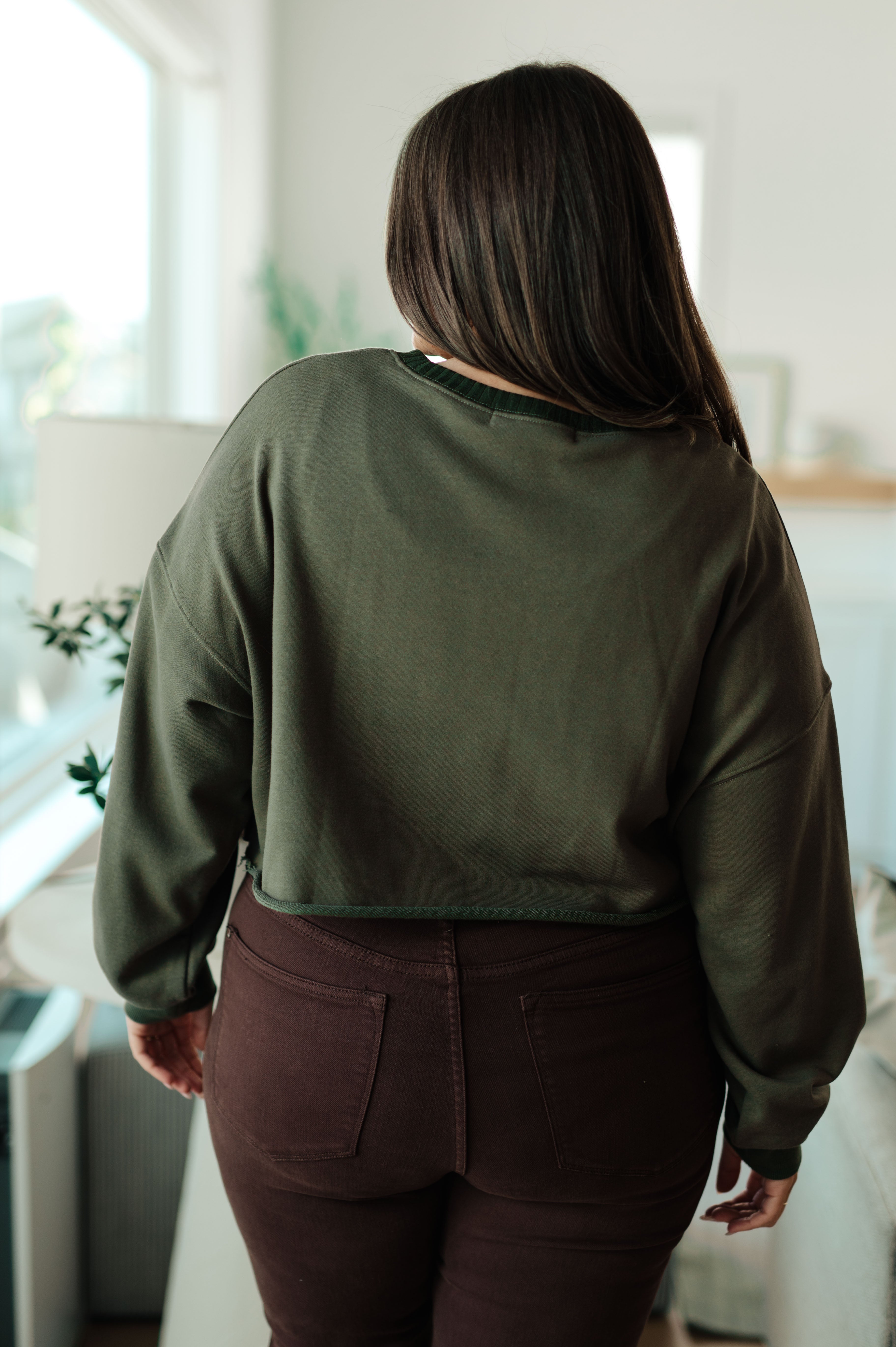 Rear view of person indoors wearing green Just a Casual Crop Pullover with long brown hair and dark pants