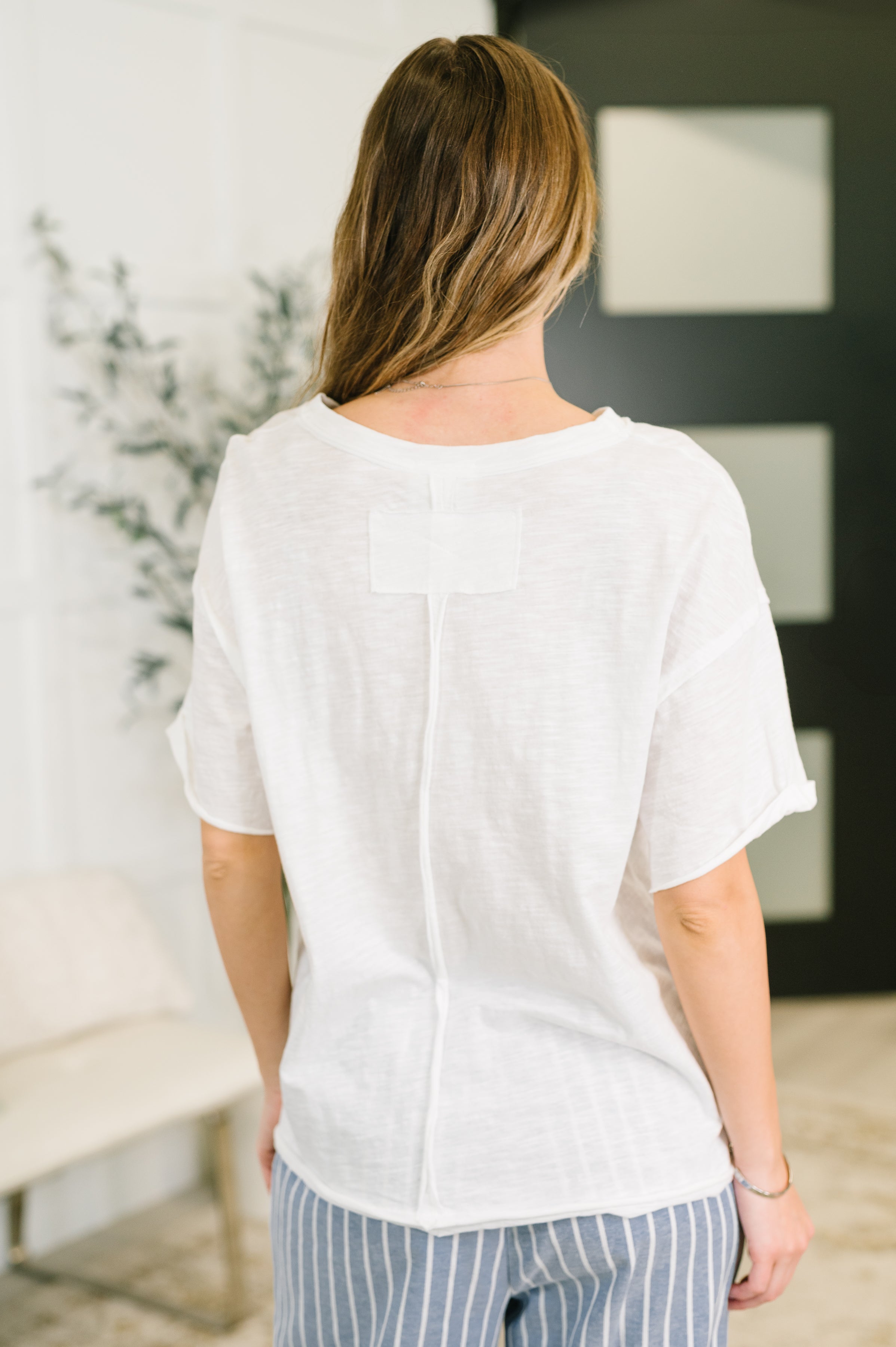 Woman seen from behind wearing an ivory relaxed tee with rolled sleeves and striped pants indoors in bright room