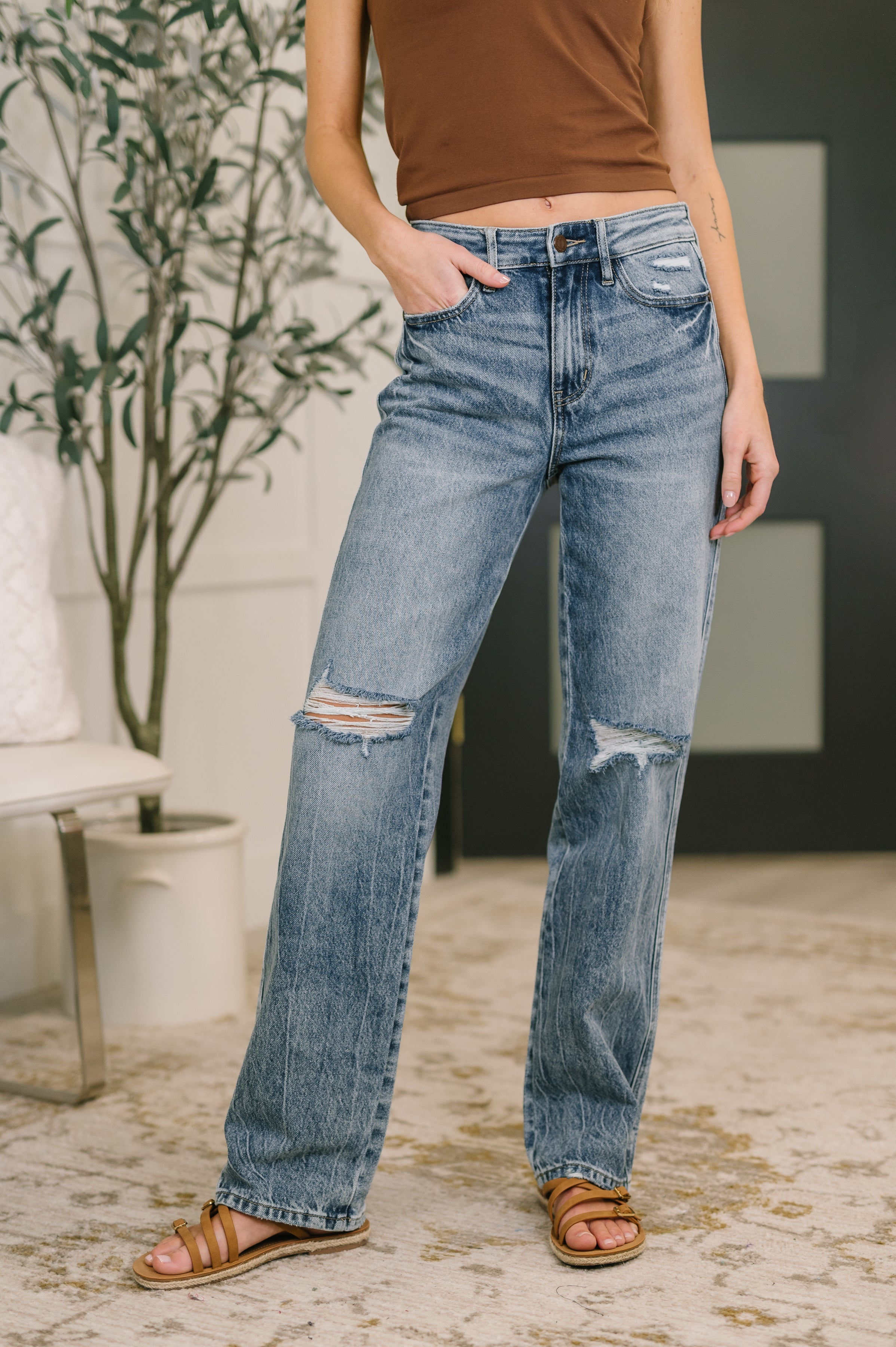 Woman wearing high waist faded blue rigid straight jeans with distressed knee rips and brown sandals indoors