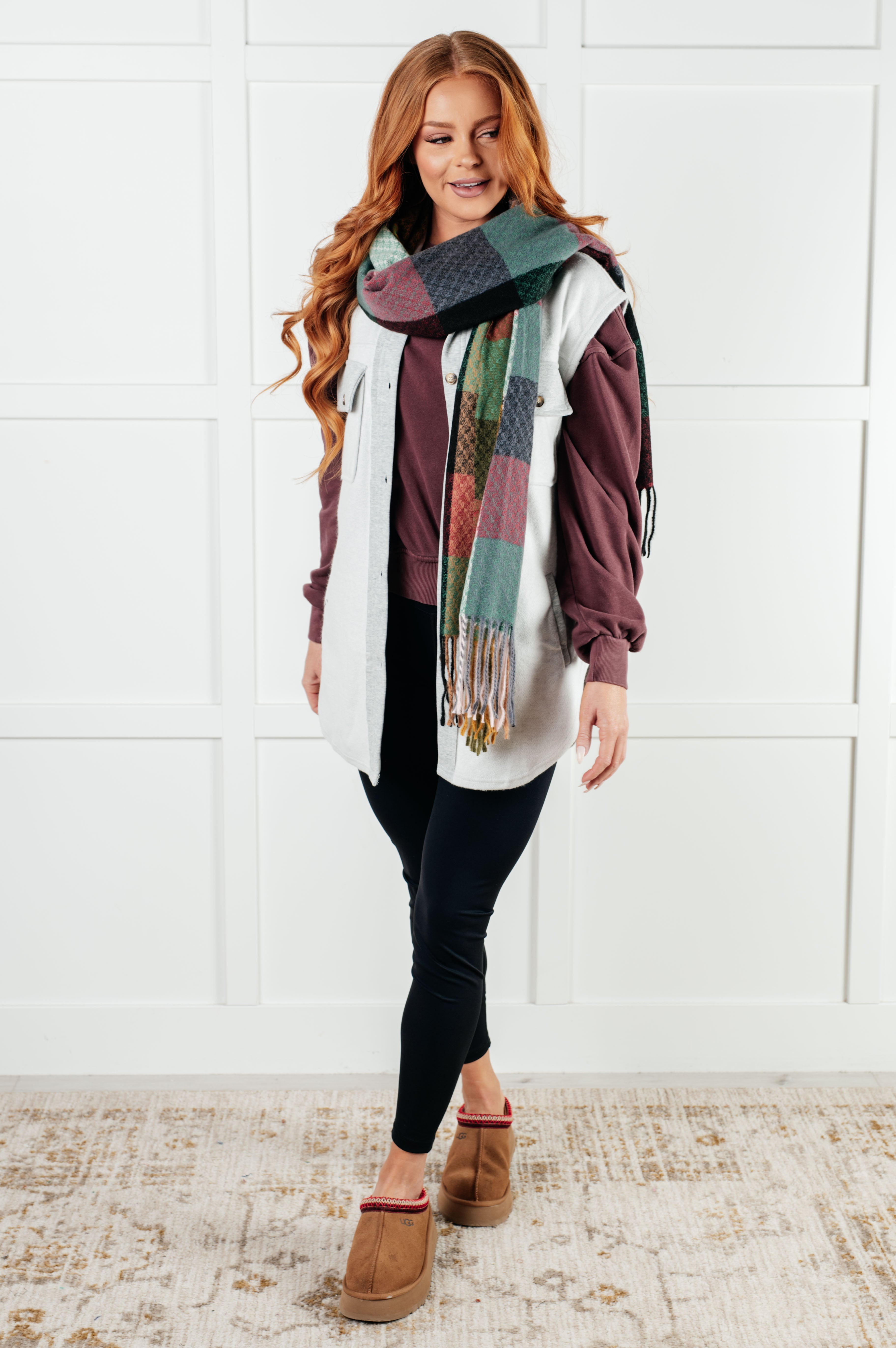 Woman wearing a colorful checker scarf with fringe, purple sweater, white vest, black pants, and brown shoes against a white wall.