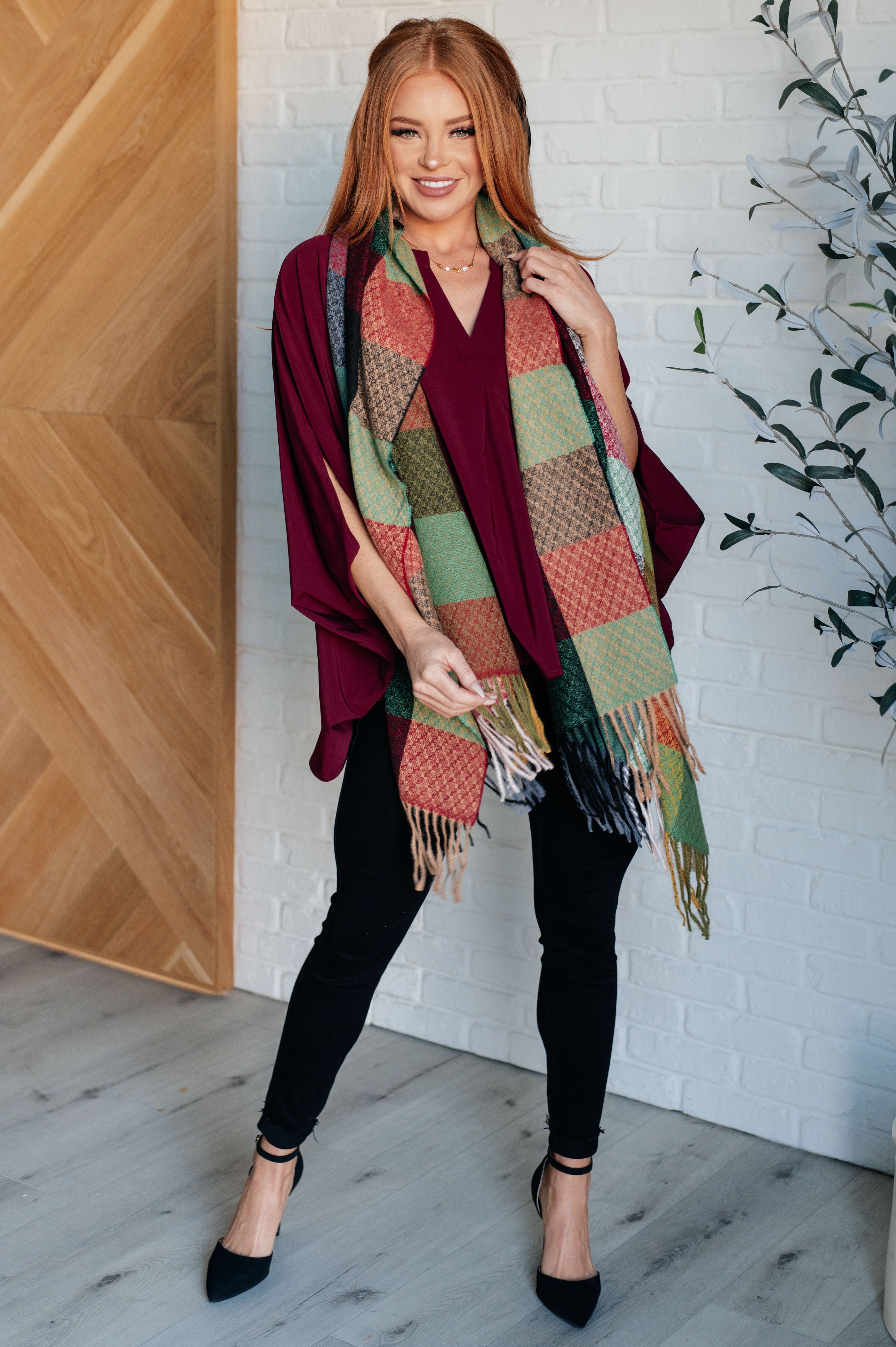 Woman wearing a colorful checkered scarf with fringe, a burgundy flowy top, black pants and black shoes indoors against a white brick wall.