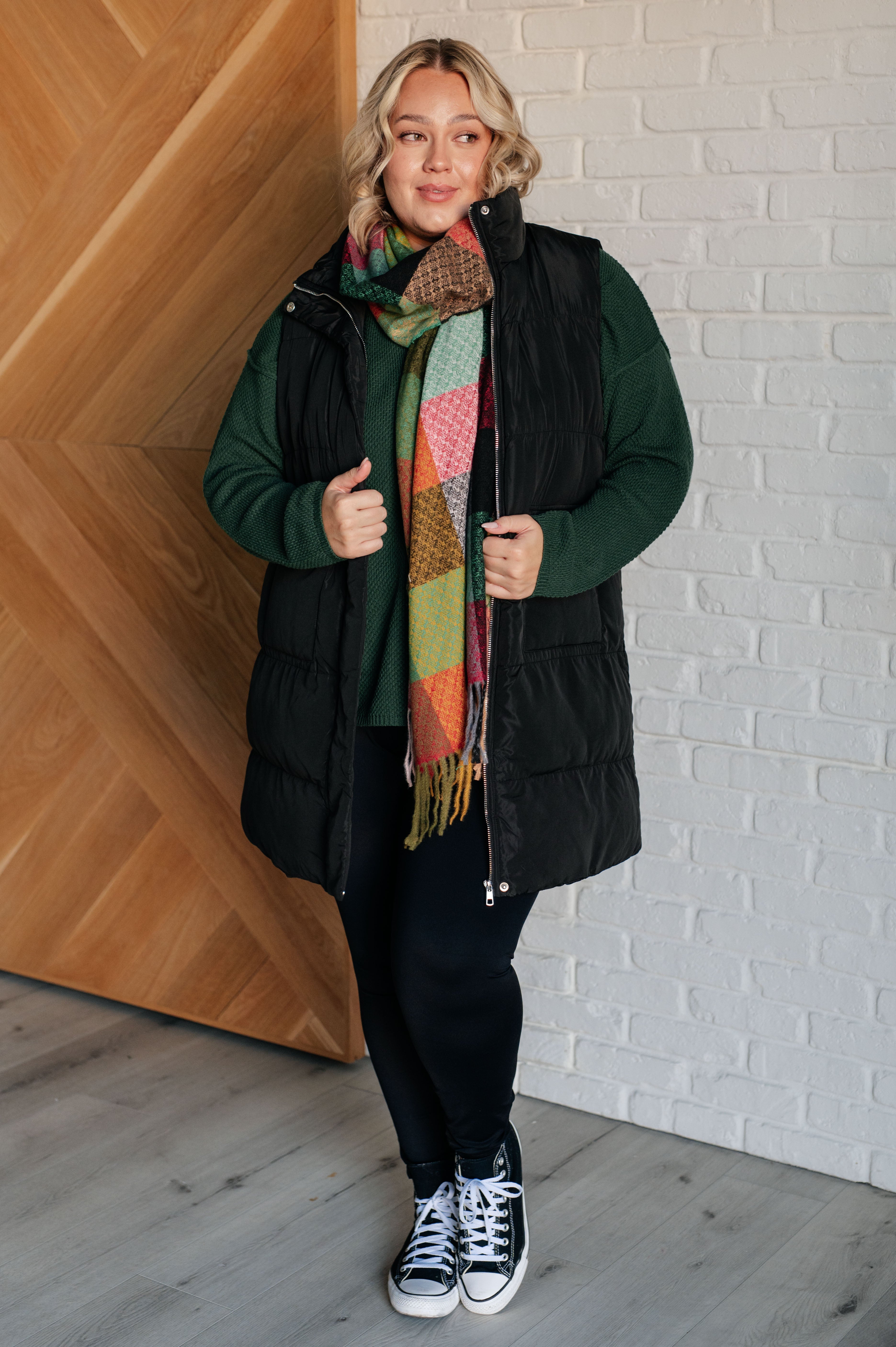 Woman wearing a black vest, green sweater, colorful checkered scarf with fringe, black pants and black converse sneakers against a white brick wall.
