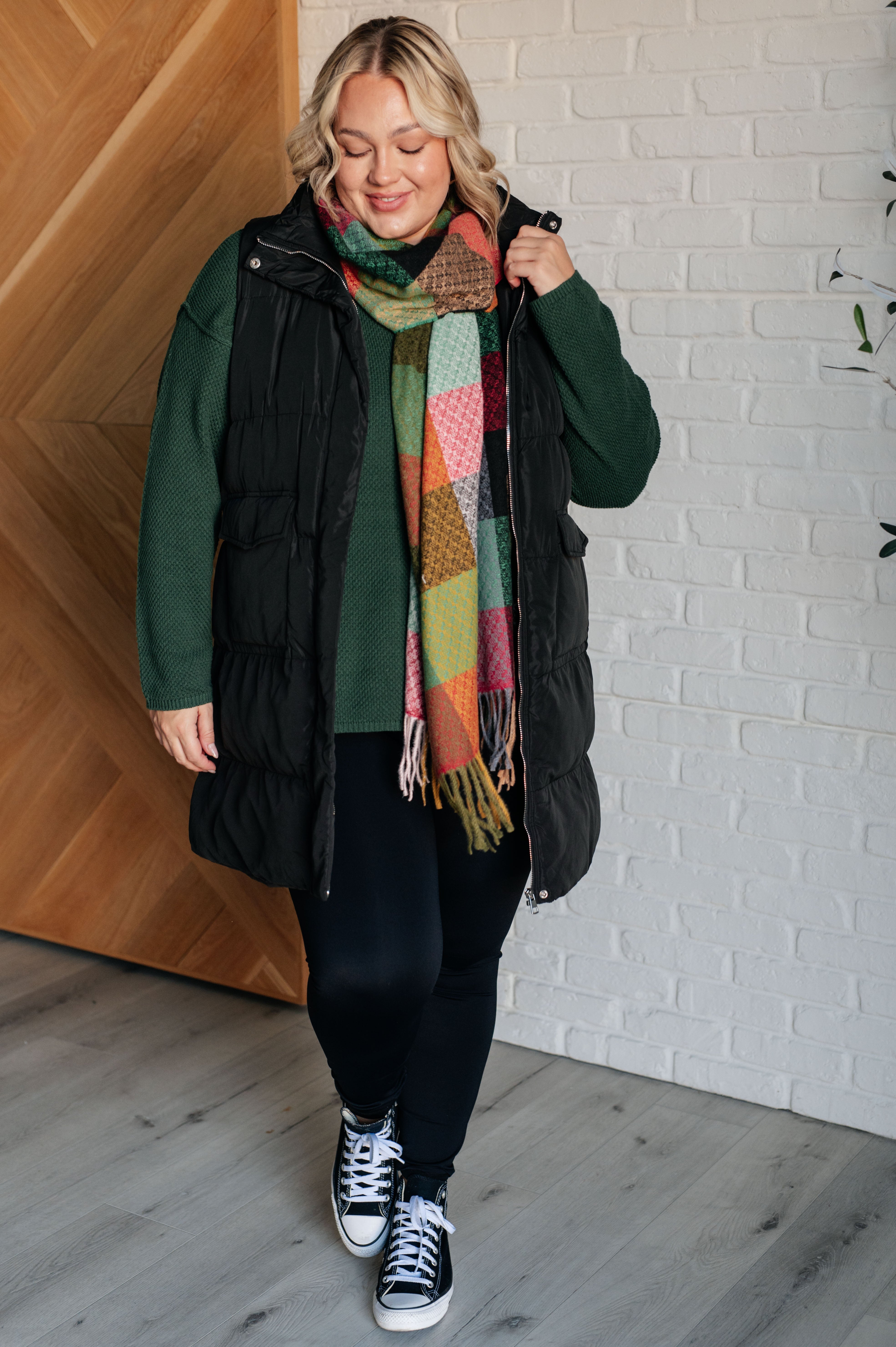 Woman wearing a green sweater, black vest, colorful checkered scarf and with fringe, black pants and black converse sneakers indoors.