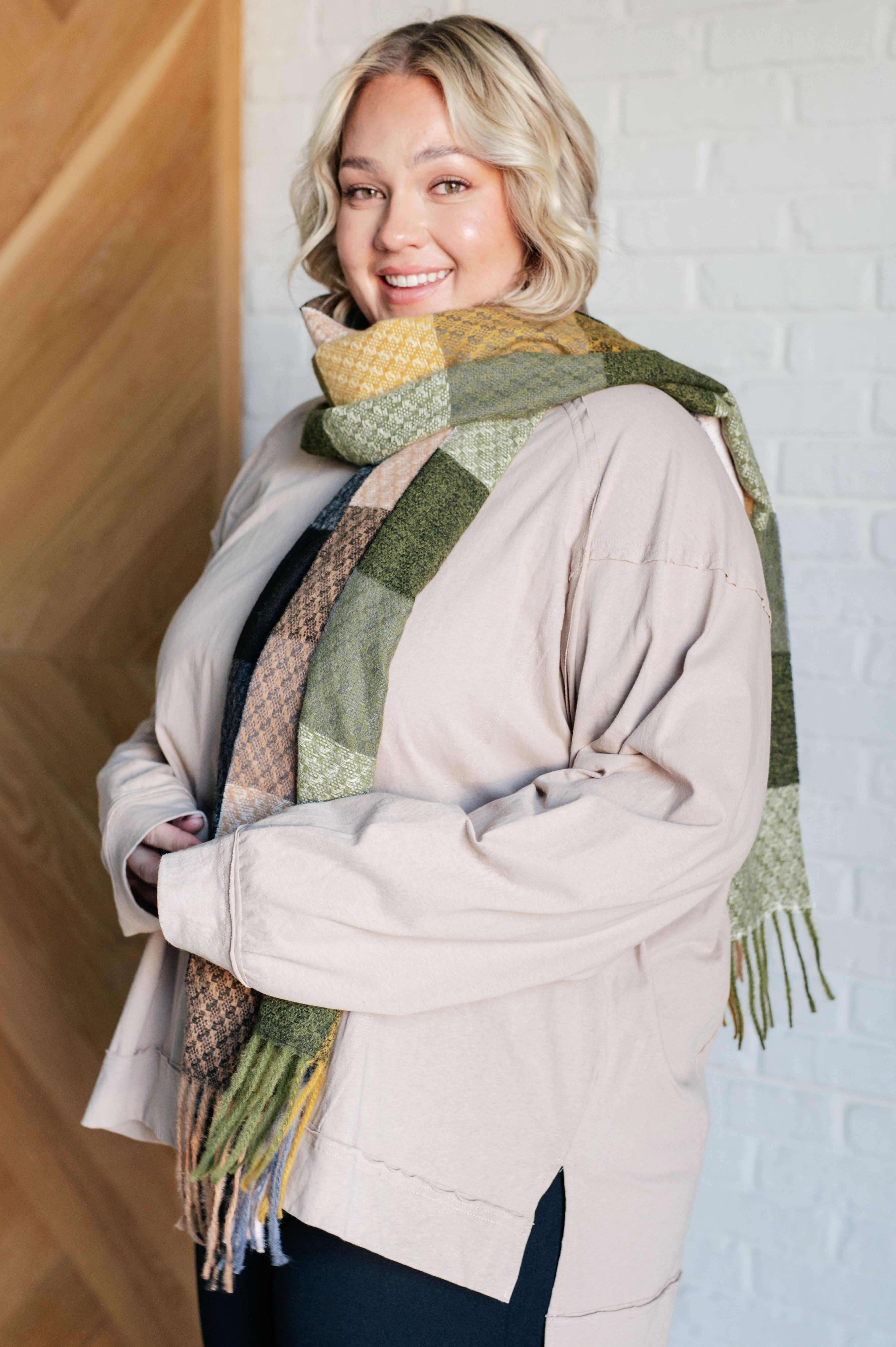 Woman wearing a large, colorful checkered scarf with fringe, a beige sweater and navy pants against a white brick wall.