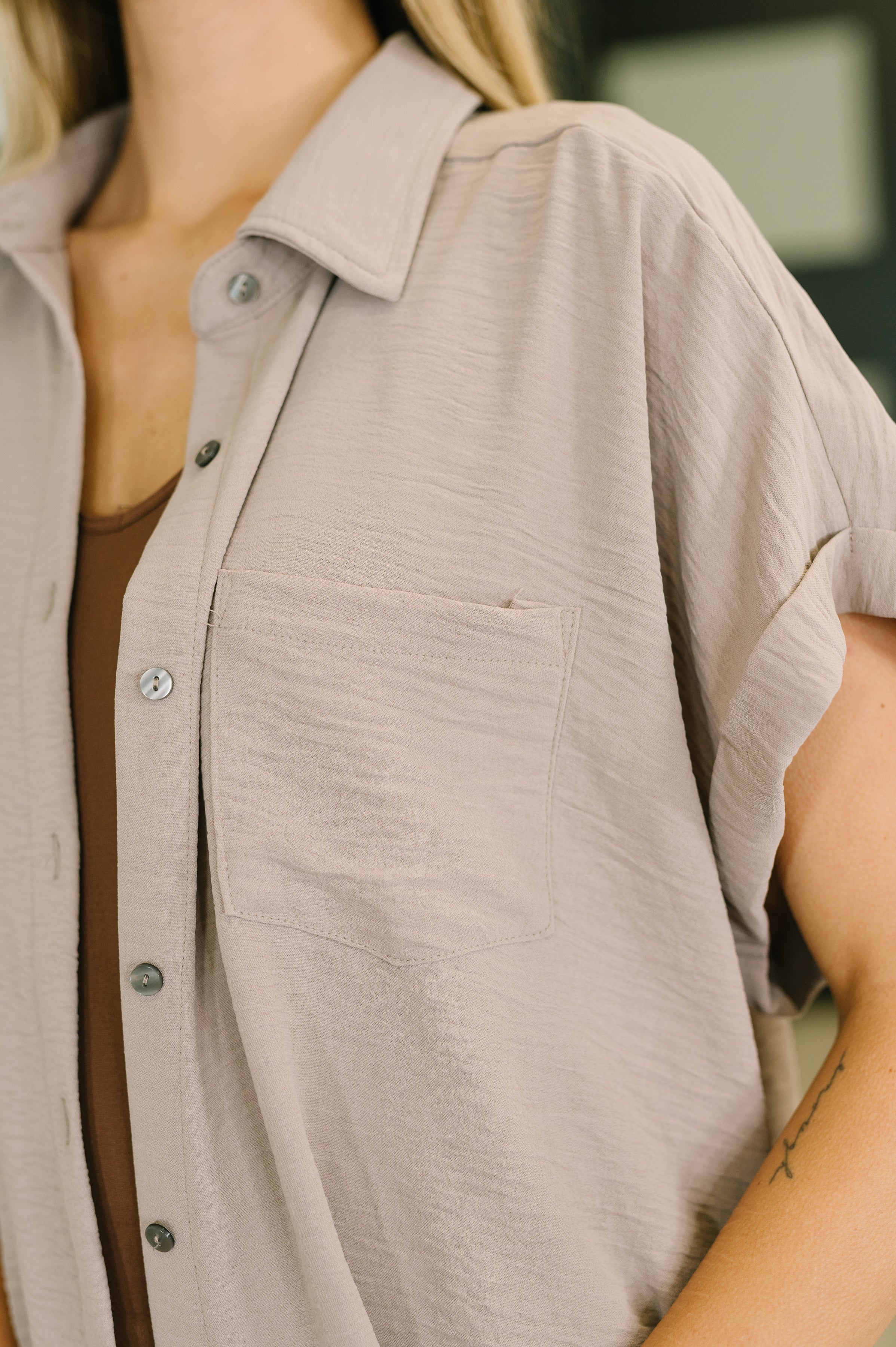 Close-up of a woman wearing ash mocha short sleeve collared button-down shirt with chest pocket over brown top