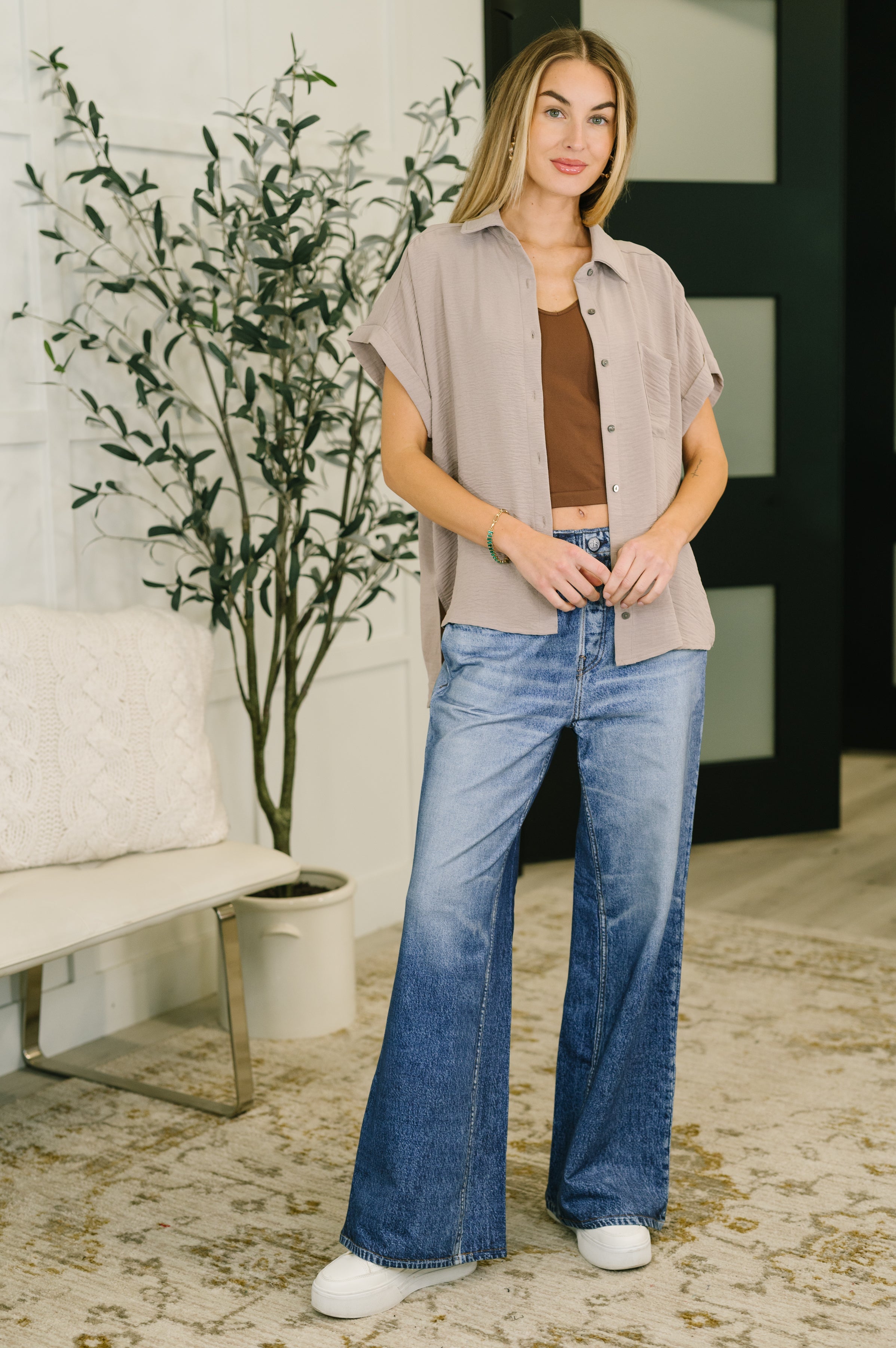 Woman wearing loose fit ash mocha short sleeve collared button down shirt with blue wide leg jeans and white sneakers indoors