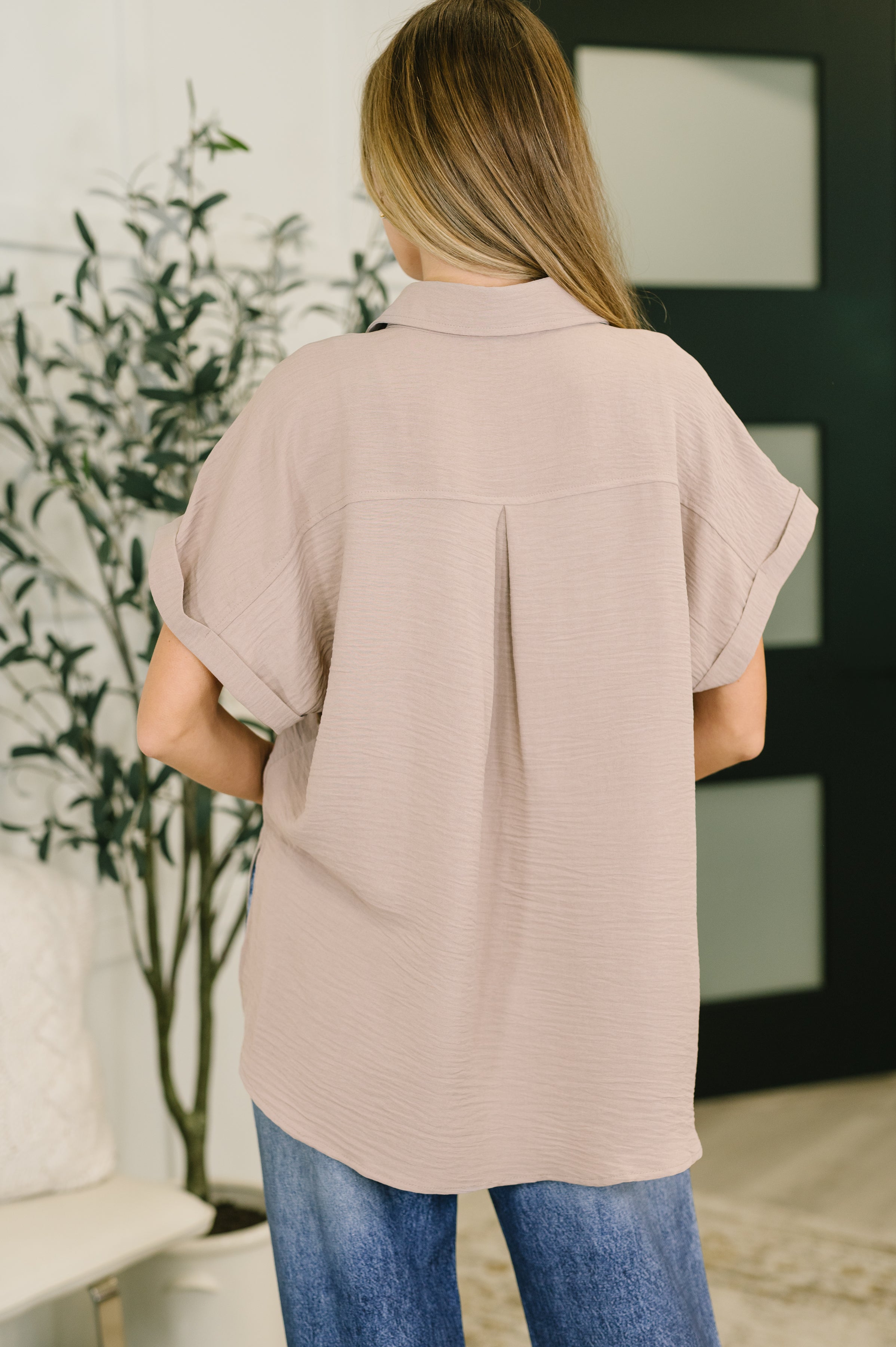 Woman with long hair wearing loose fit ash mocha collared button down shirt and blue jeans standing indoors