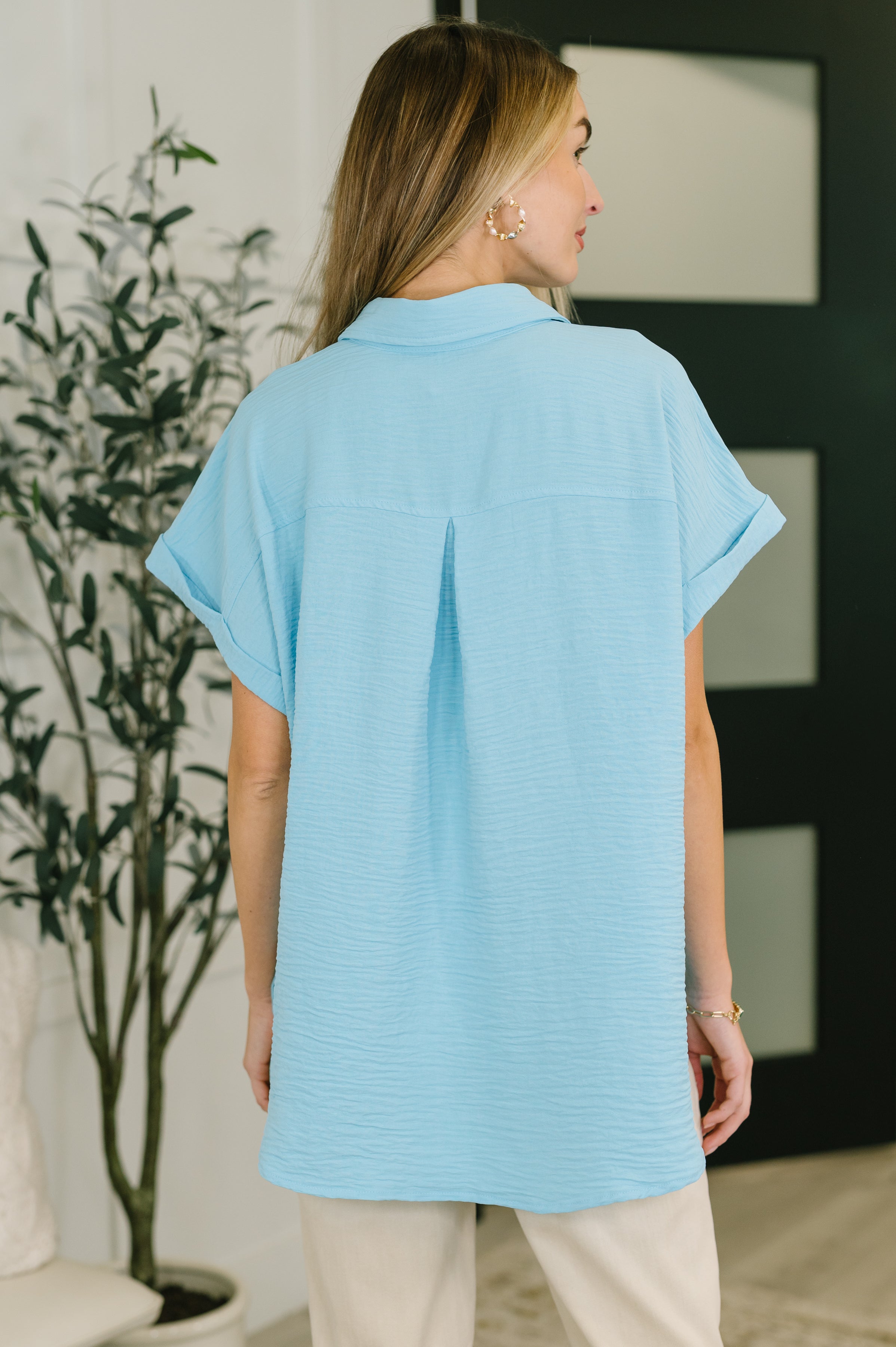 Woman wearing loose fit sky blue short sleeve collared button down shirt with cuffed sleeves seen from the back indoors