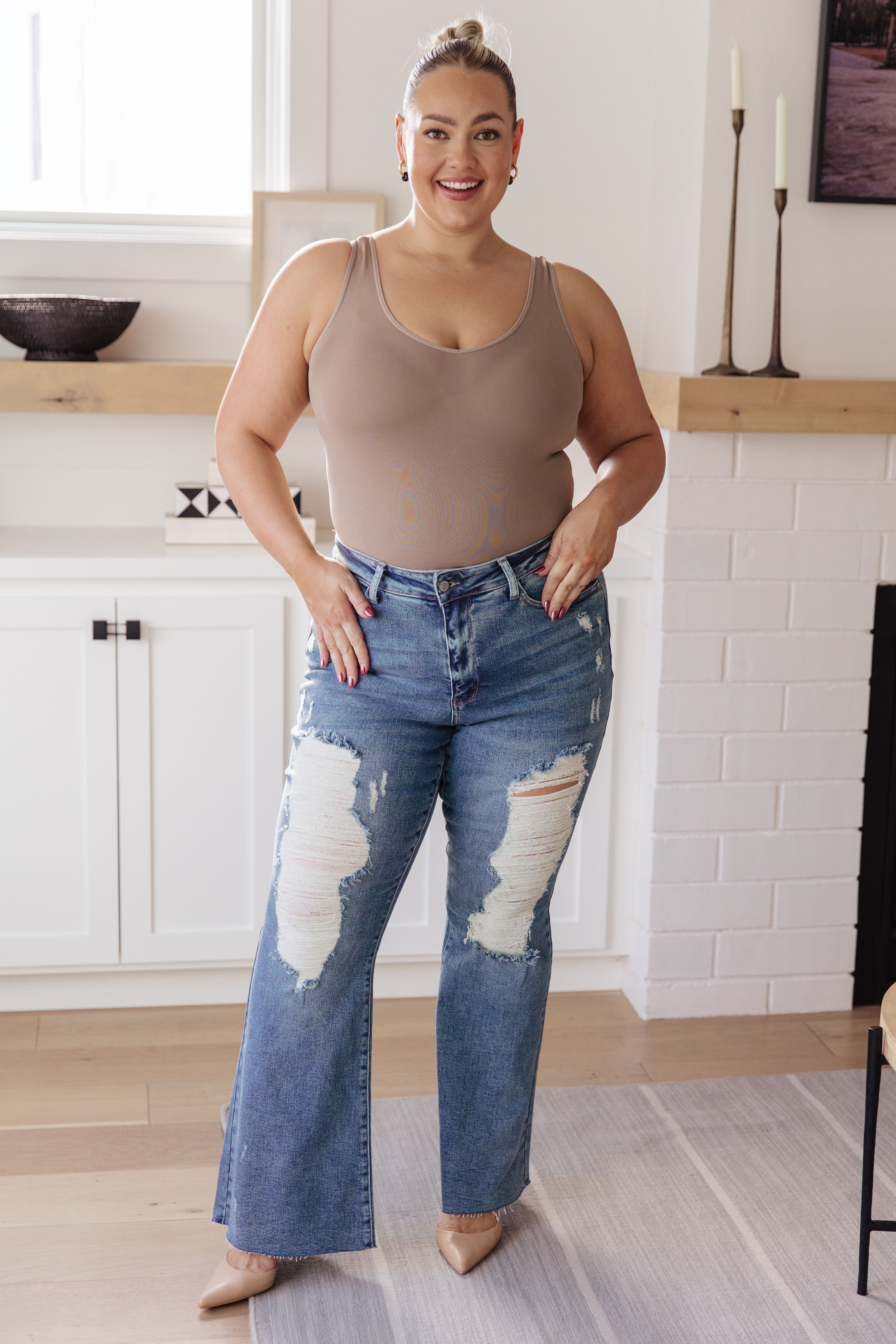 Plus-size woman wearing light brown sleeveless top with high-rise blue ripped flare jeans indoors