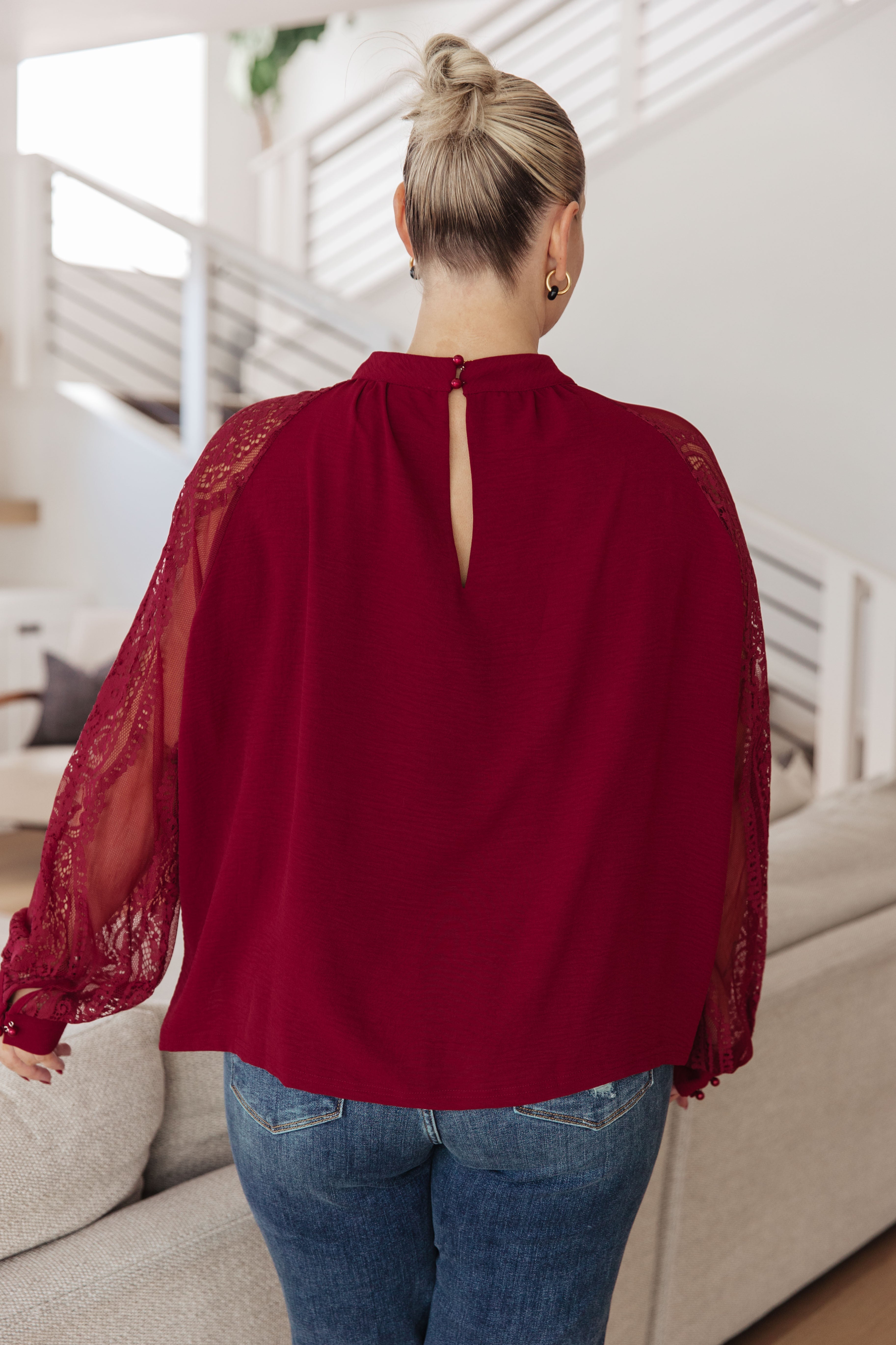 Back view of person wearing wine blouse with lace bishop sleeves and keyhole button closure at the neckline indoors