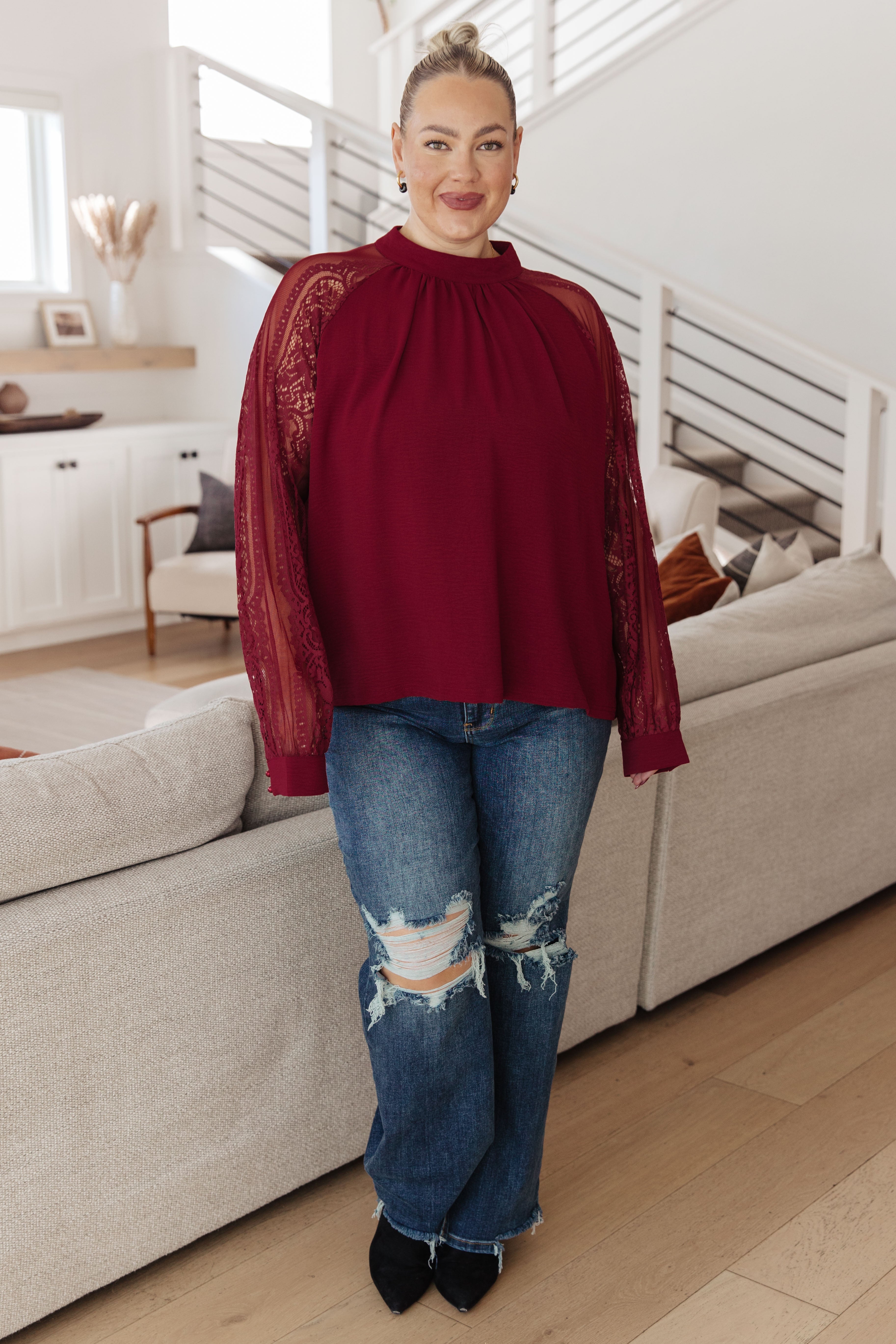 Woman wearing a wine-colored blouse with lace long sleeves and gathered neckline paired with distressed jeans indoors