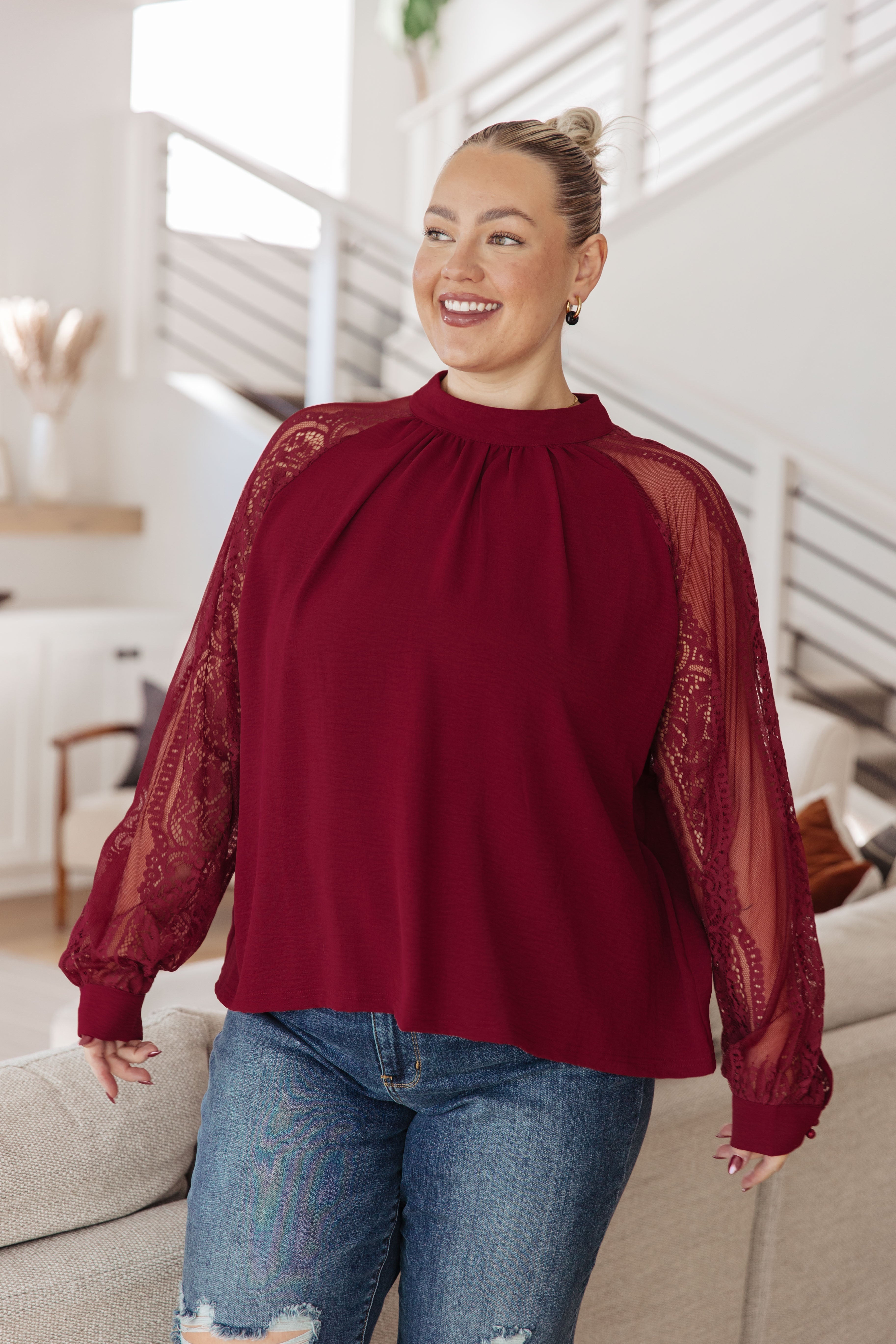 Smiling woman wearing a wine-colored blouse with sheer lace sleeves and gathered neckline paired with ripped blue jeans indoors