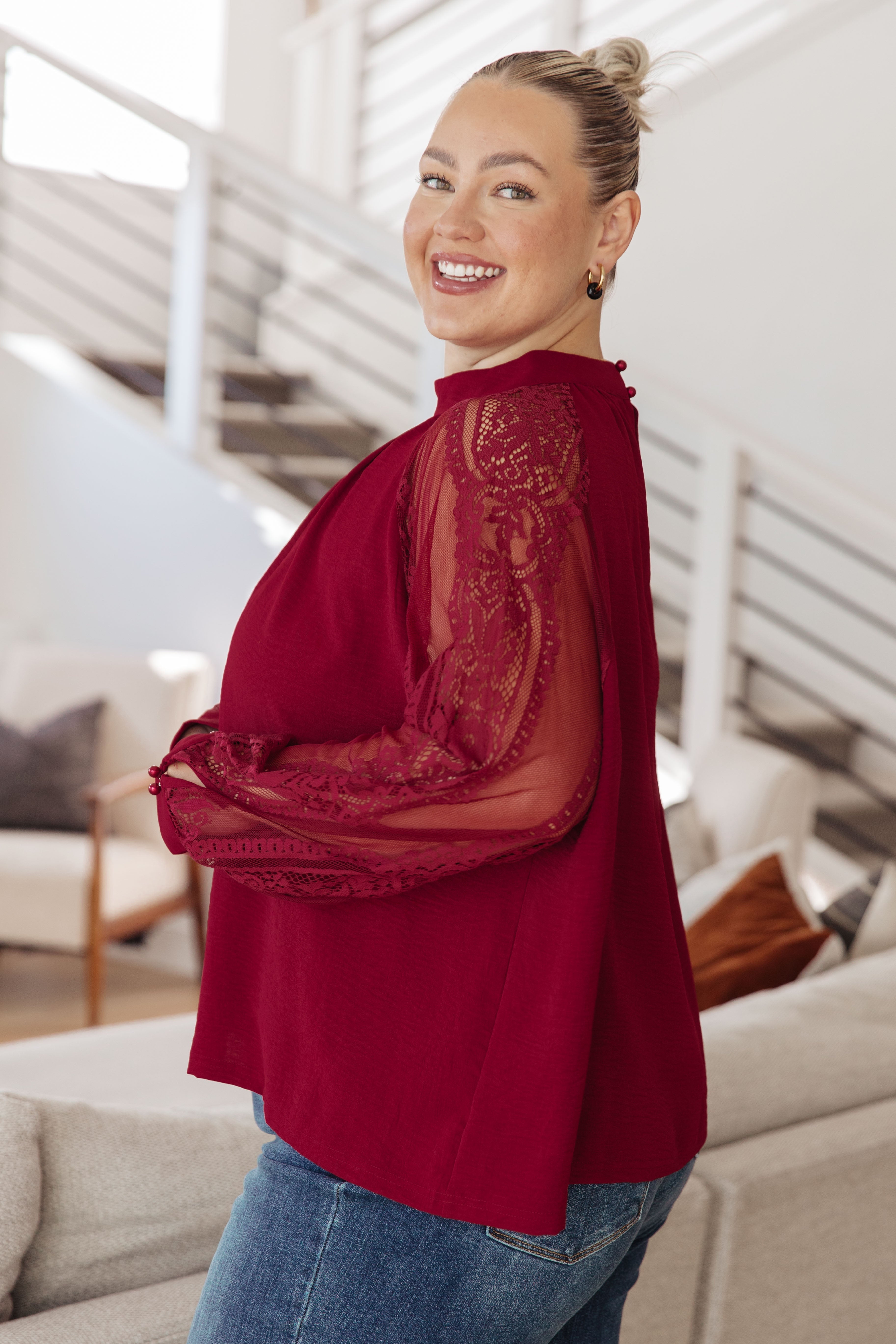 Smiling woman wearing a rich wine blouse with sheer lace sleeves and buttoned cuffs standing indoors in casual jeans