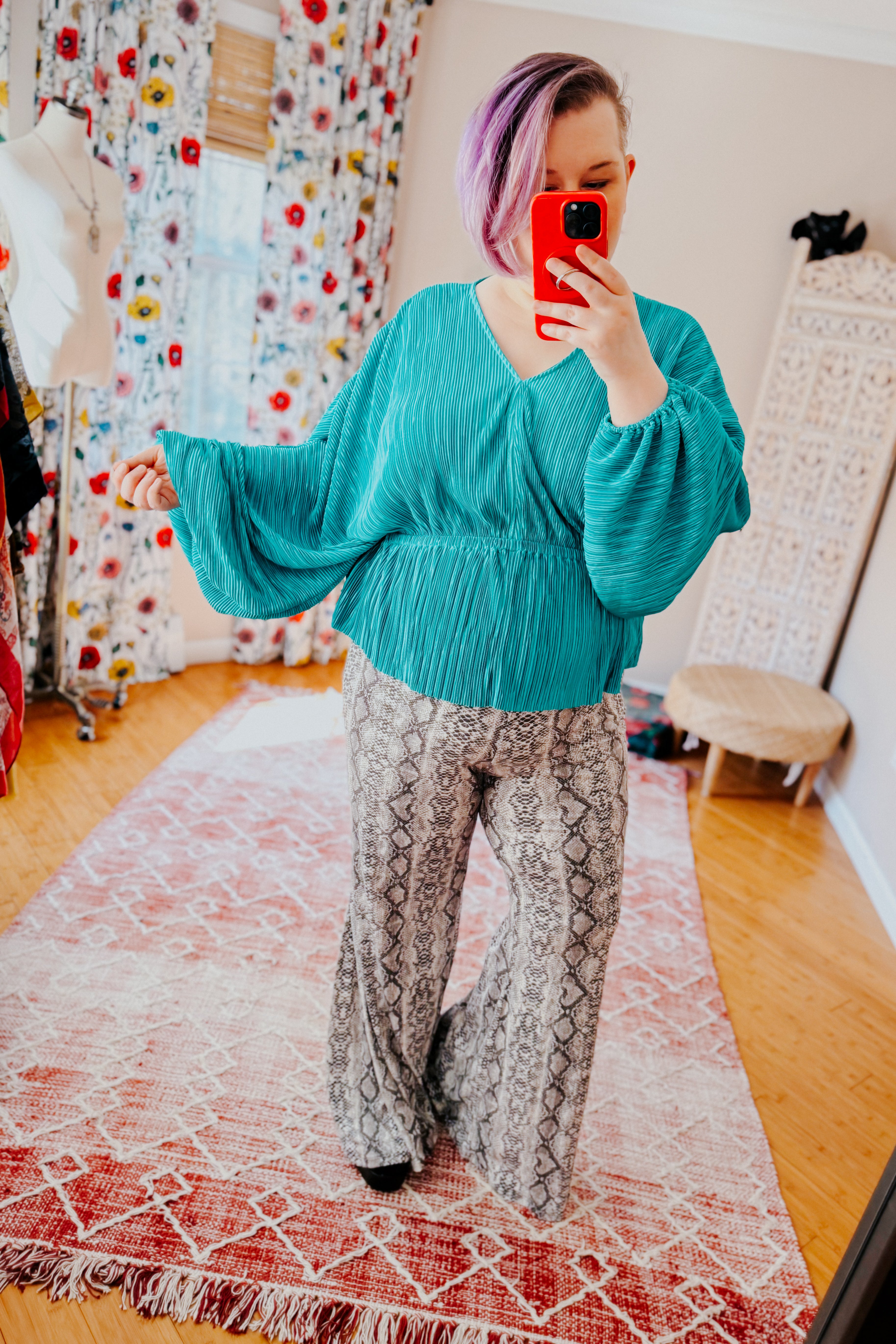 Woman with short purple hair wearing a teal pleated top with wide sleeves and snake print wide-leg pants indoors