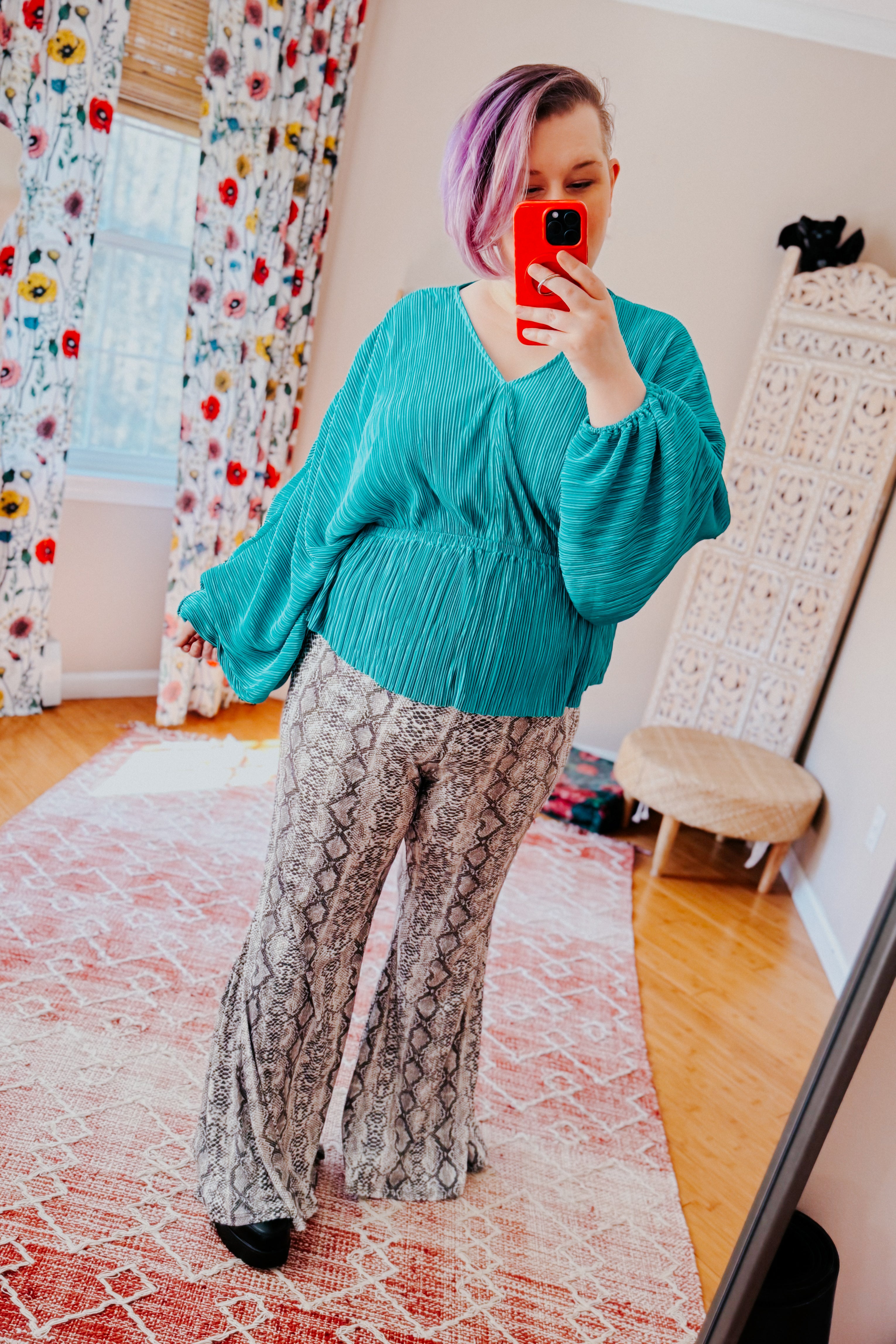 Woman wearing teal textured wrap blouse with voluminous sleeves and snake print flared pants taking mirror selfie indoors