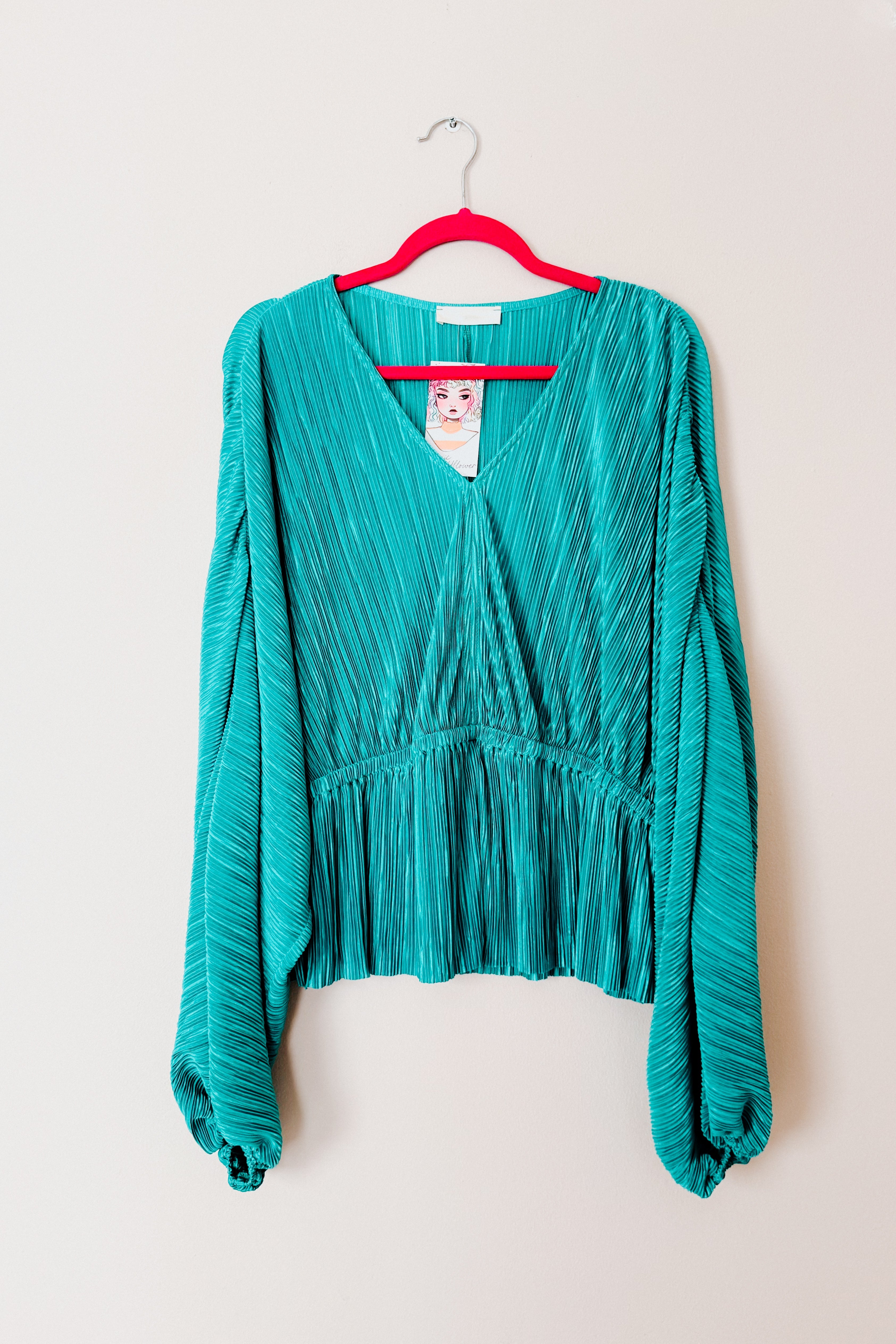 Teal pleated blouse with deep V-neck and voluminous bishop sleeves hanging on a bright pink hanger against a white background