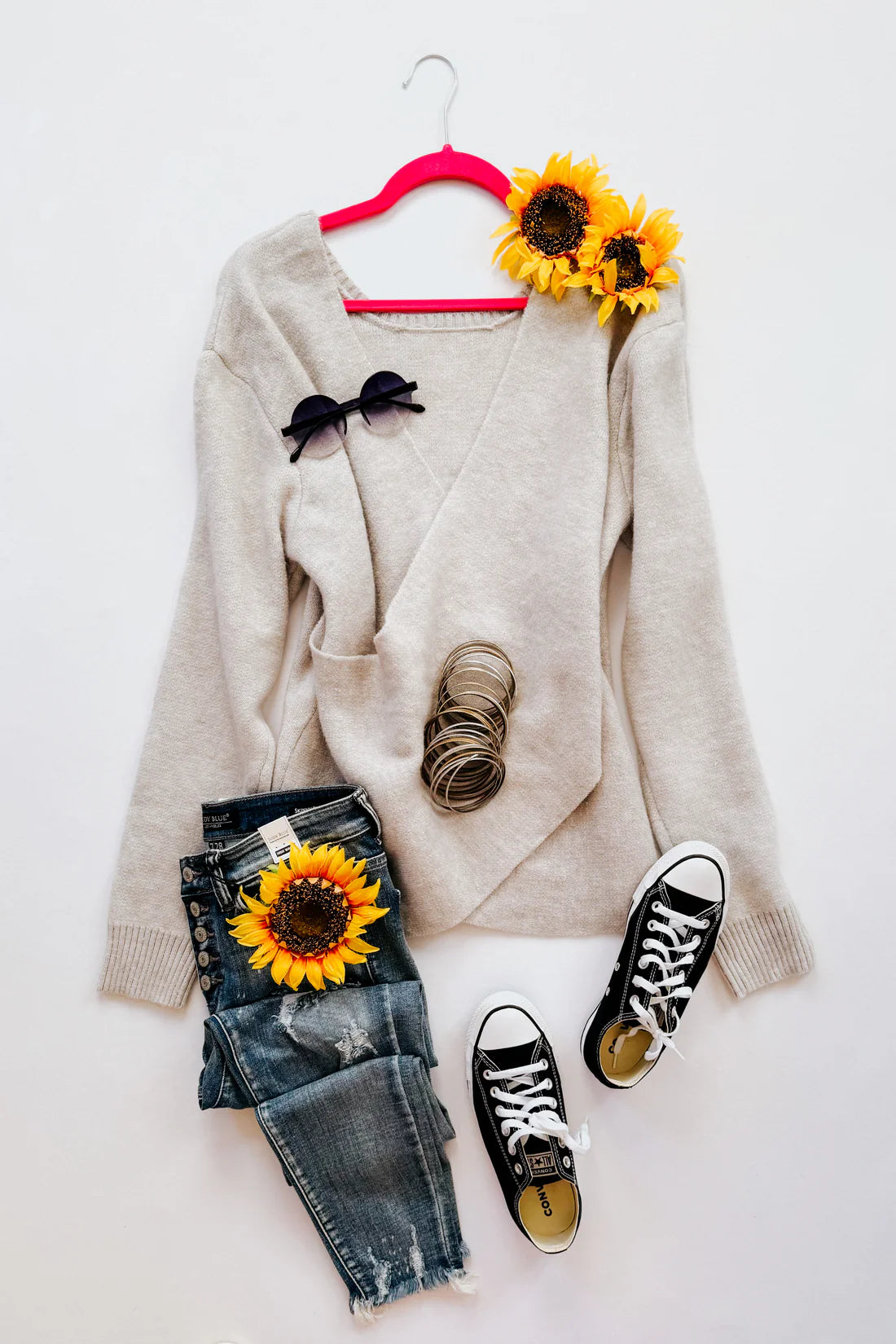 Light grey wrap sweater with ribbed cuffs styled with distressed denim jeans, black sneakers, sunglasses, bracelets, and sunflowers on a white background
