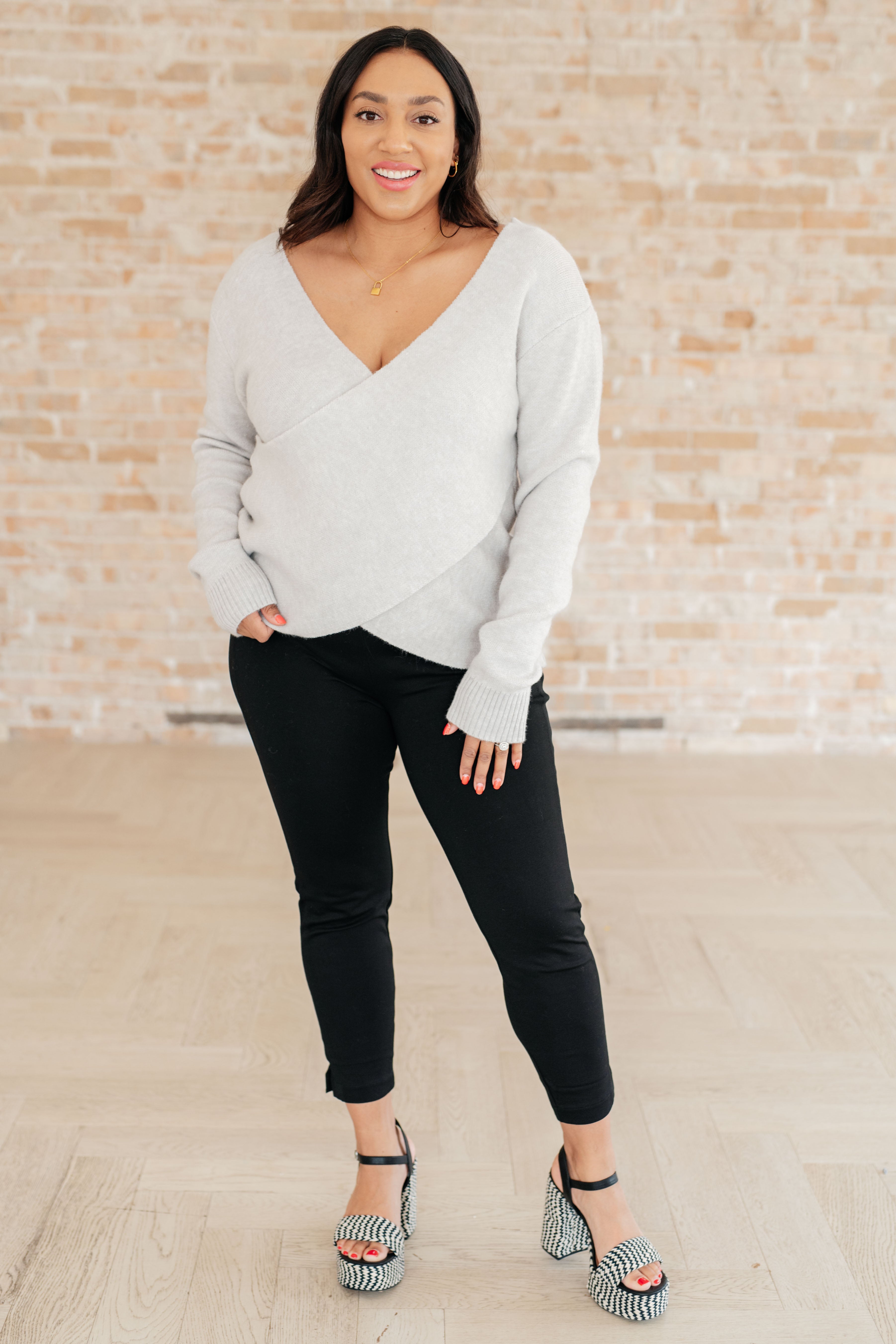 Woman wearing a grey wrap-style long-sleeve top with ribbed cuffs paired with black pants and patterned heels indoors
