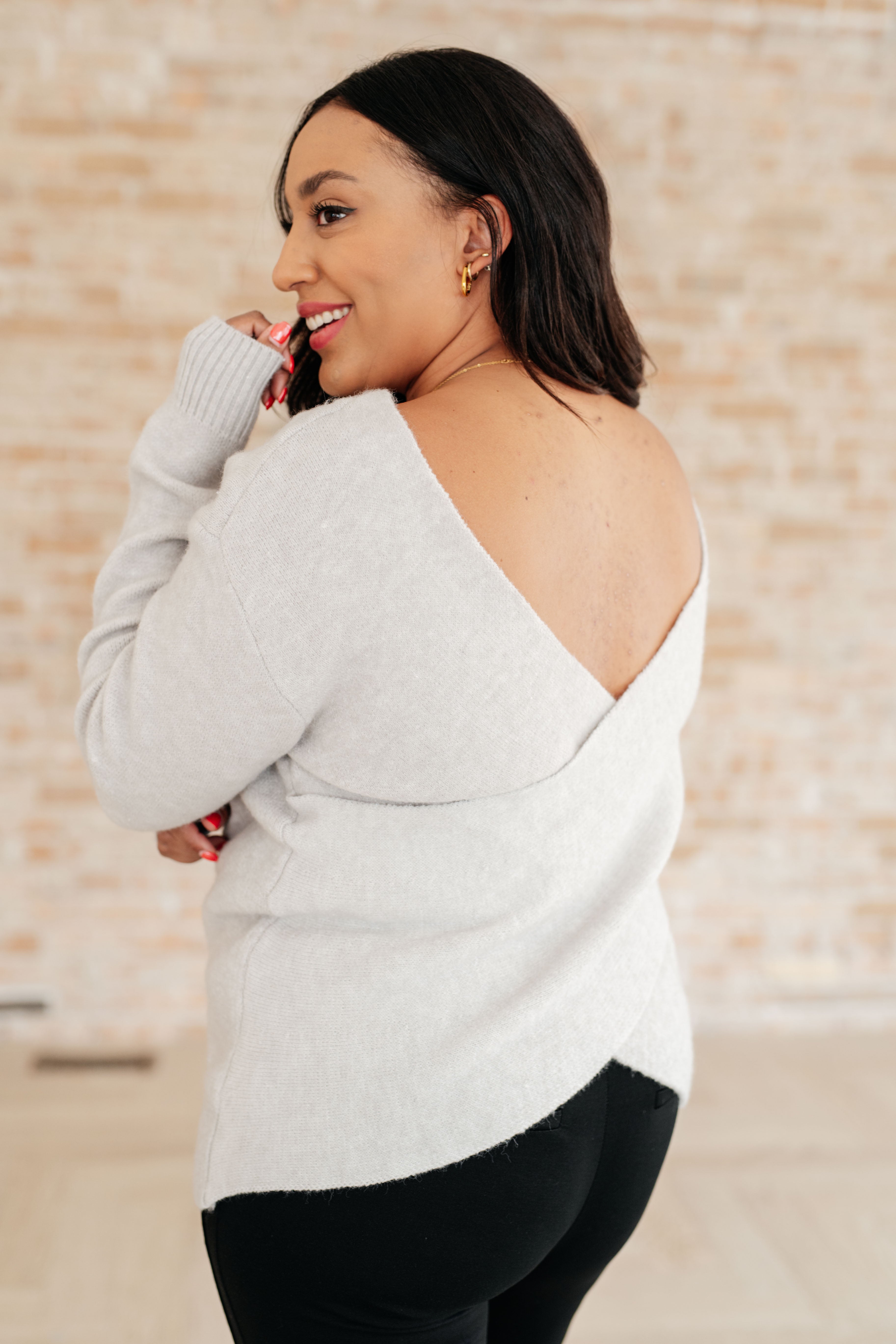 Woman smiling wearing a grey knit top with a deep V-back and ribbed cuffs paired with black pants indoors