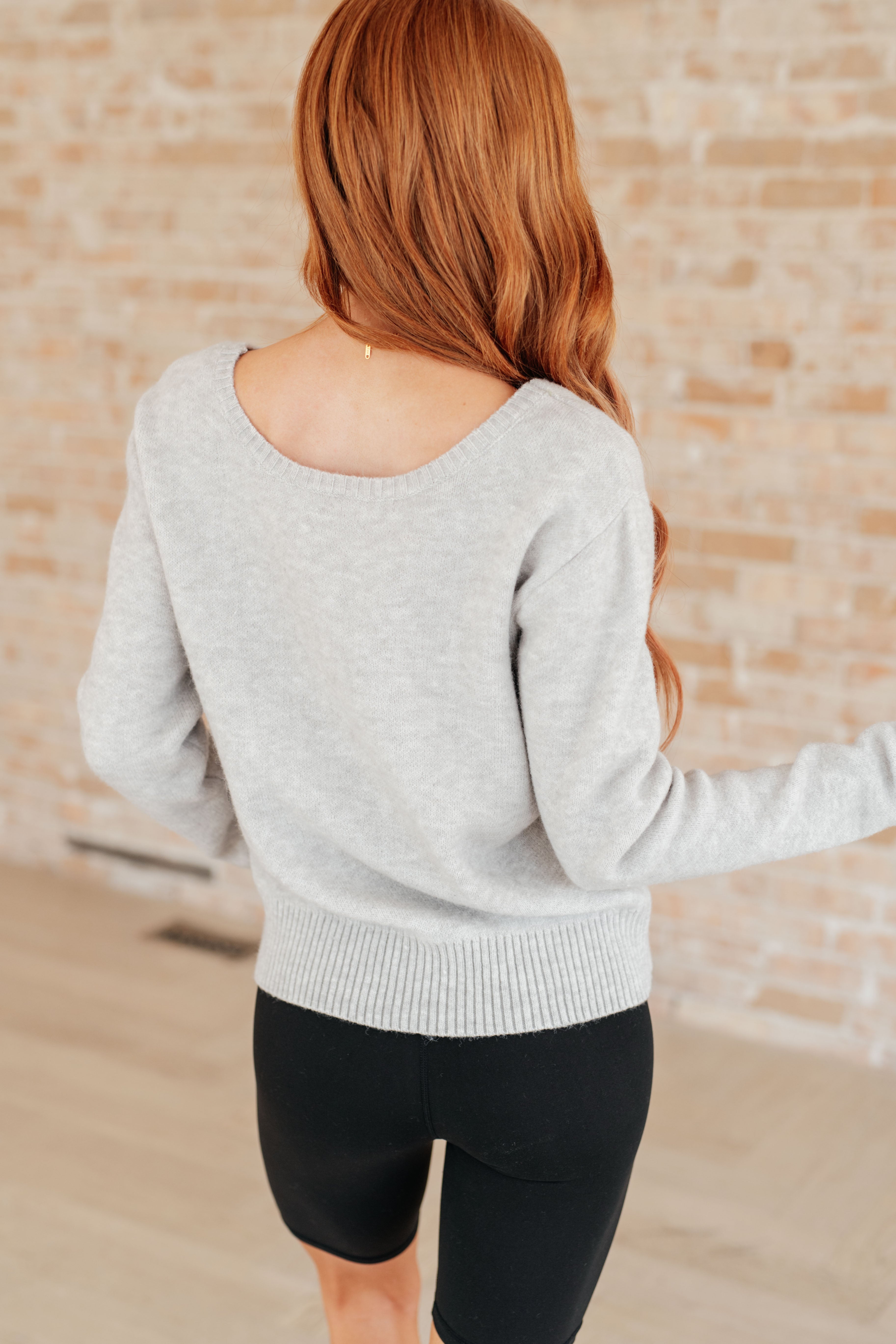 Woman with long red hair wearing a light grey long-sleeve knit top with ribbed hem and black fitted shorts indoors