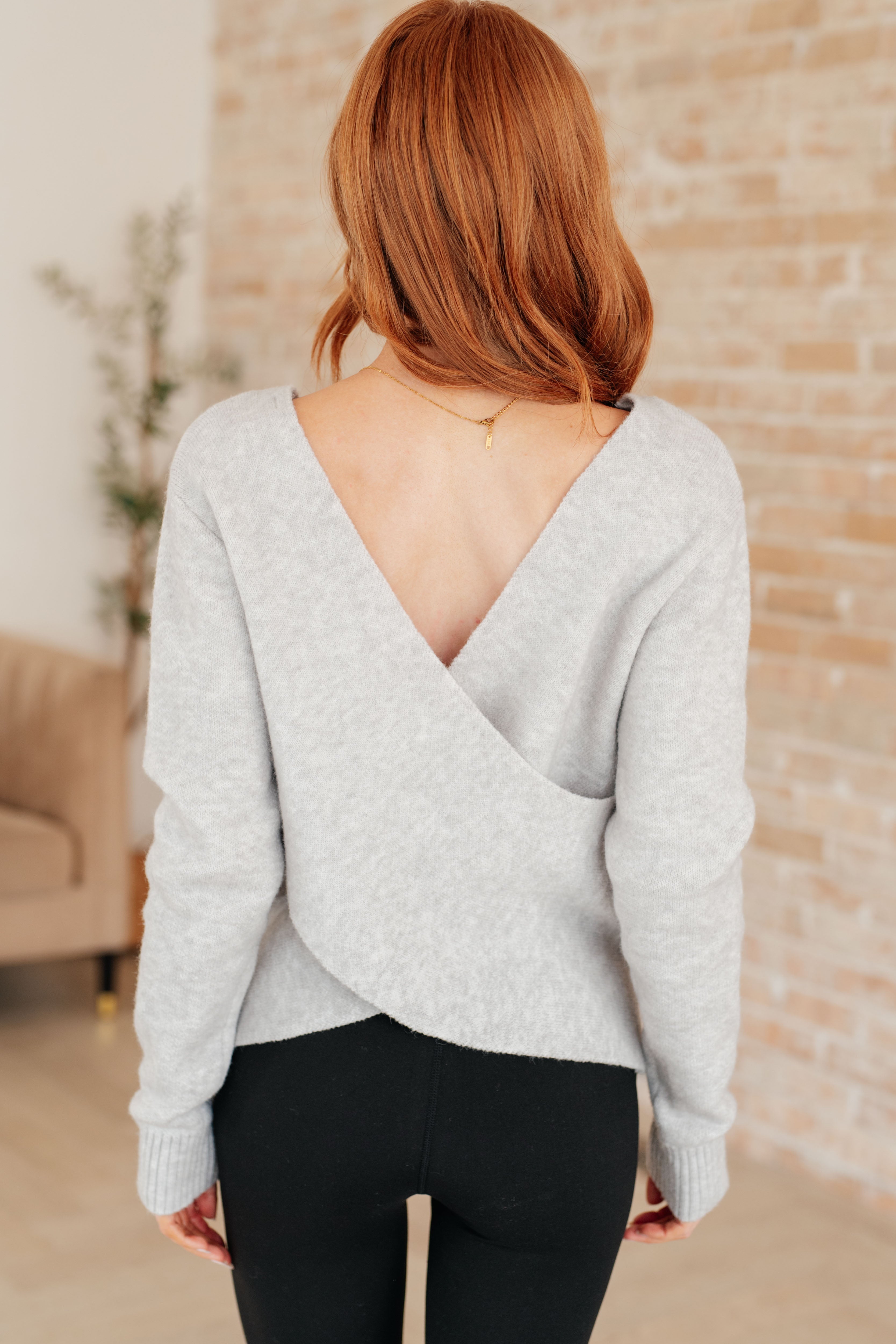 Woman with red hair wearing a light grey long-sleeve top with a deep crossover open back and black pants indoors