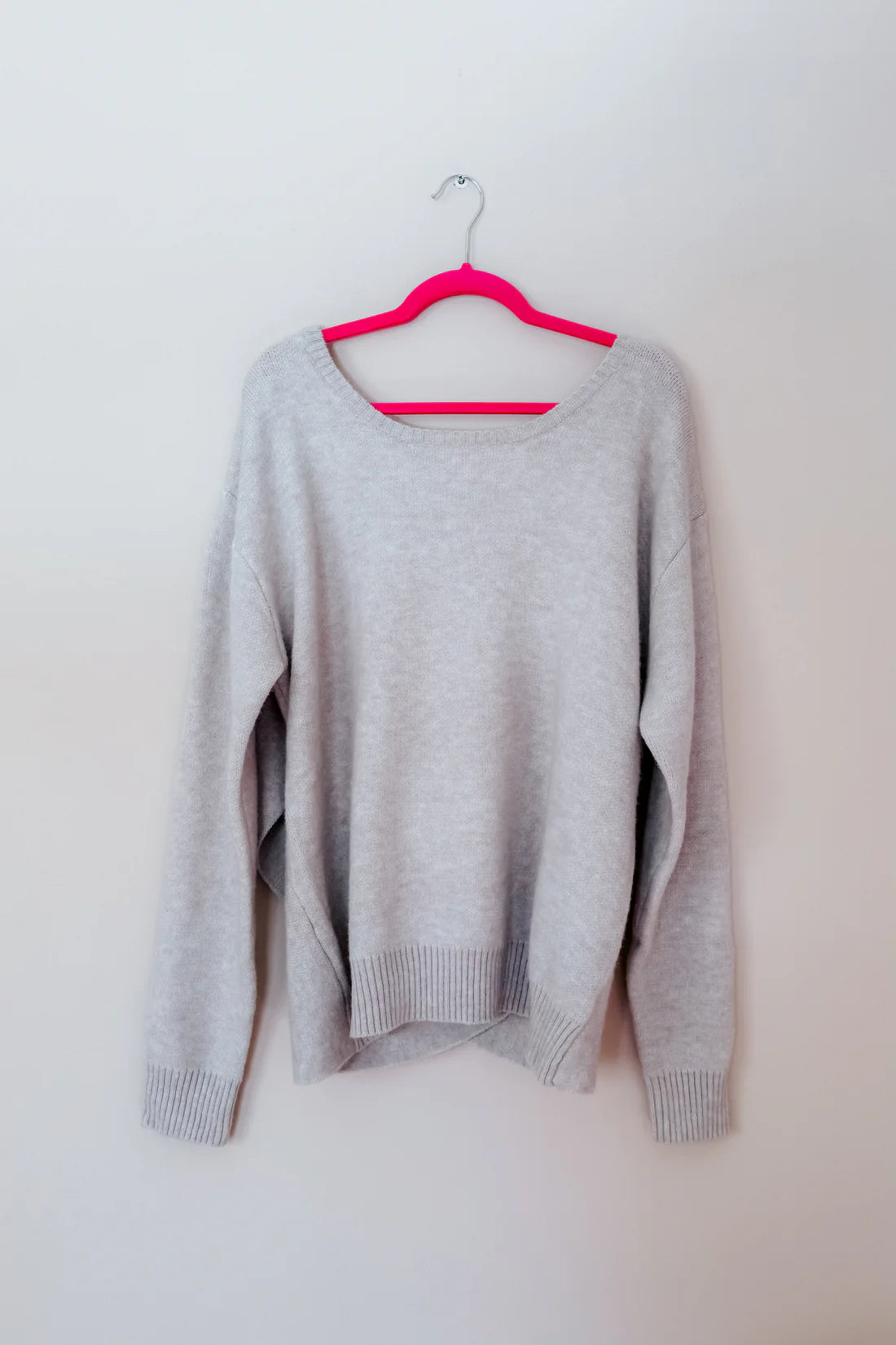 Light grey knit sweater with ribbed cuffs and hem hanging on a bright pink hanger against a plain background