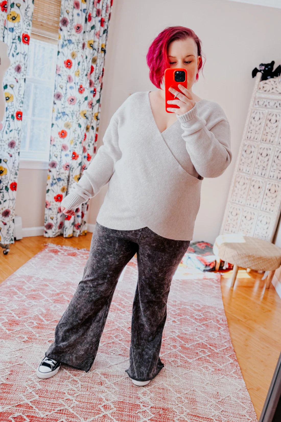 Woman with short magenta hair wearing a cozy grey wrap-style sweater paired with washed black wide-leg pants indoors