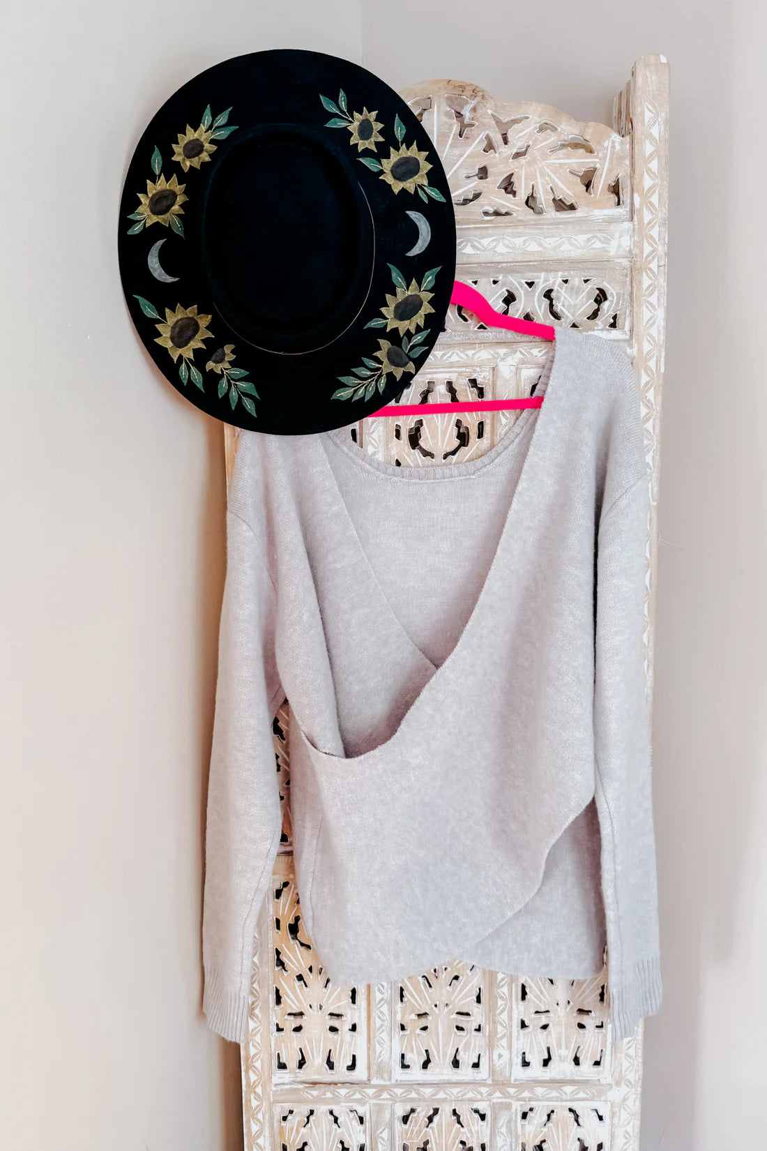 Light grey knit long-sleeve sweater with draped front detail hanging on a carved wooden panel with black floral hat