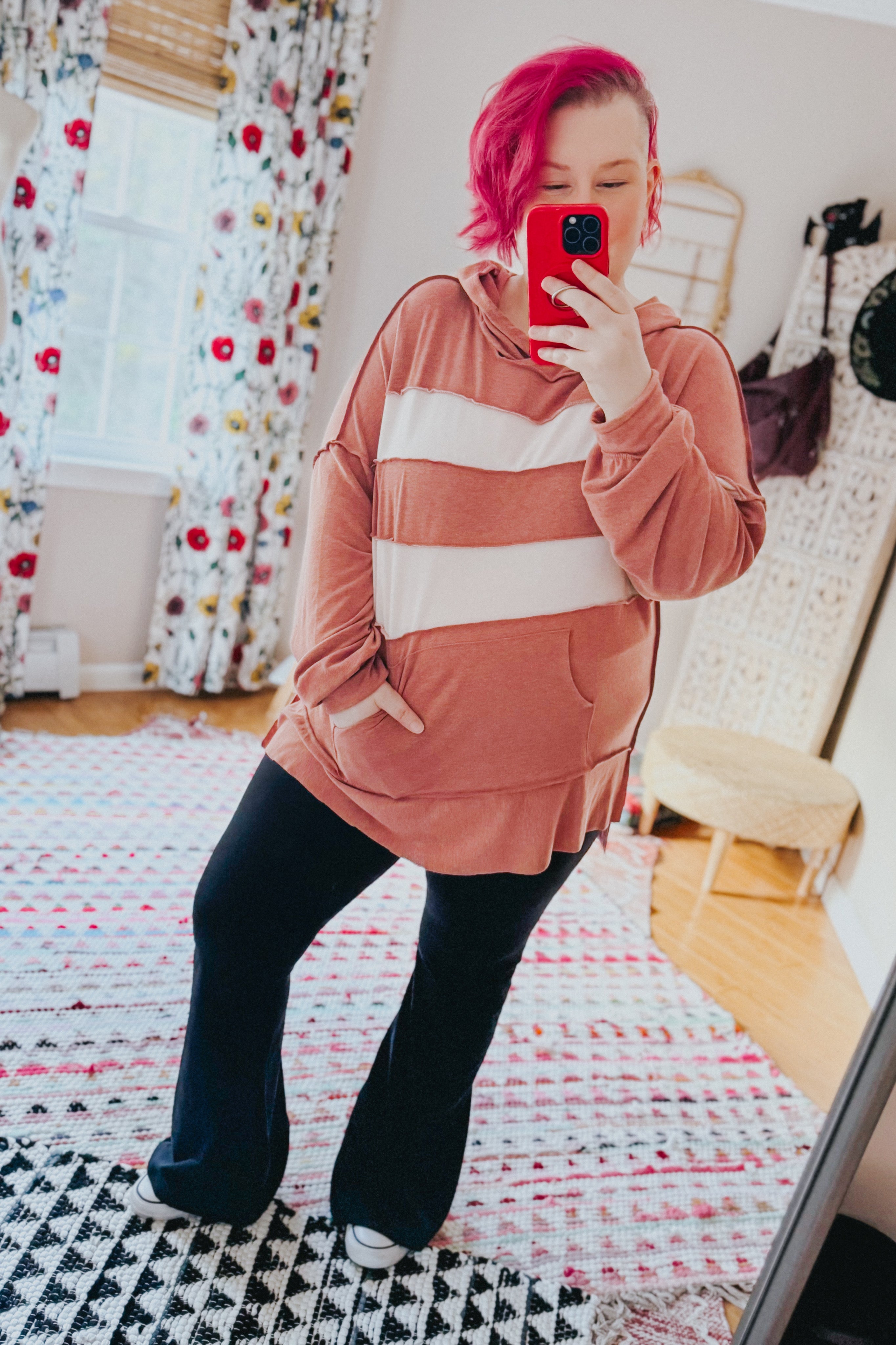 Woman with short pink hair wearing a rust-colored hoodie with white wide horizontal stripes and black pants taking a mirror selfie indoors