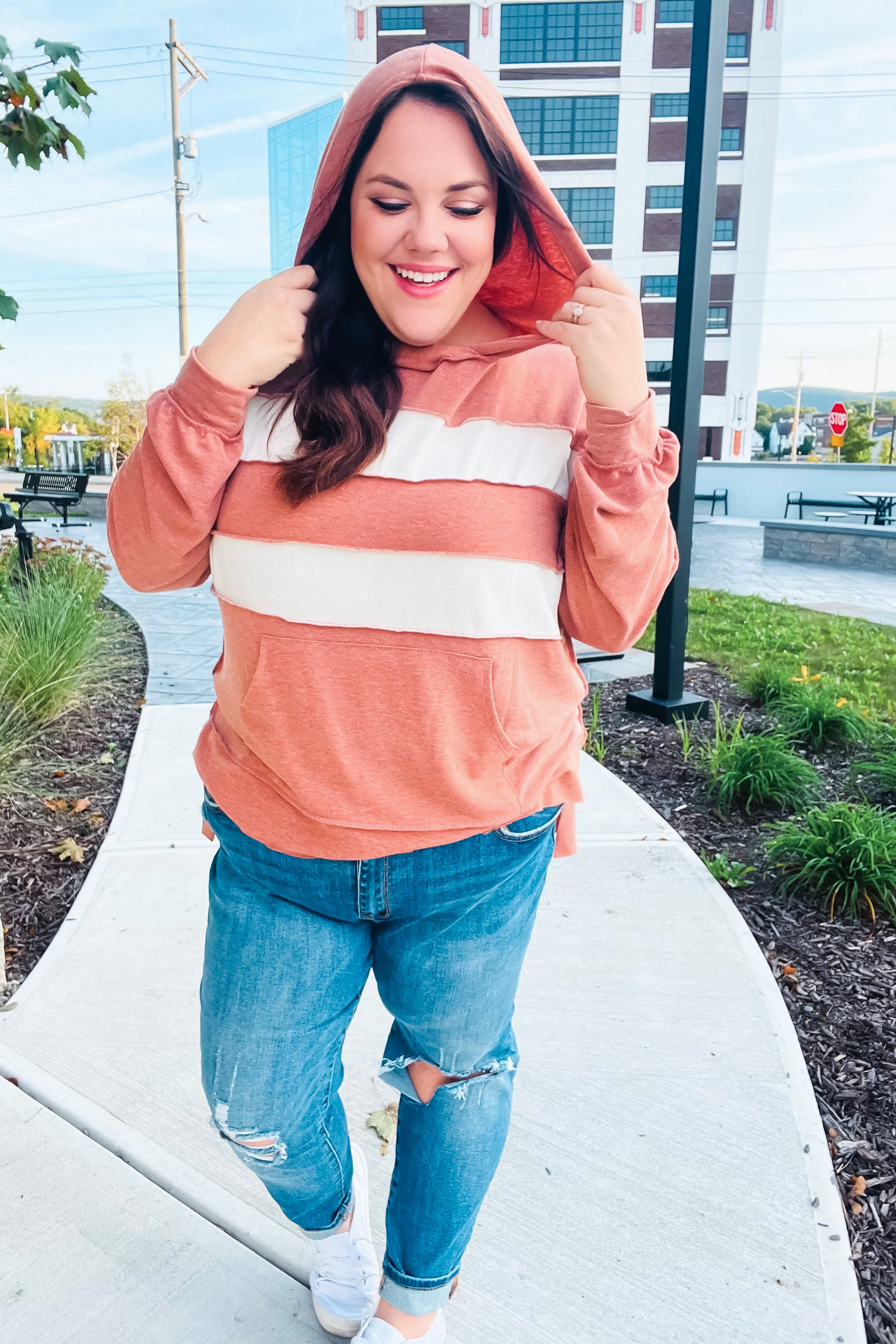 Smiling woman wearing a rust hoodie with two wide cream horizontal stripes and ripped jeans walking outdoors on a sidewalk