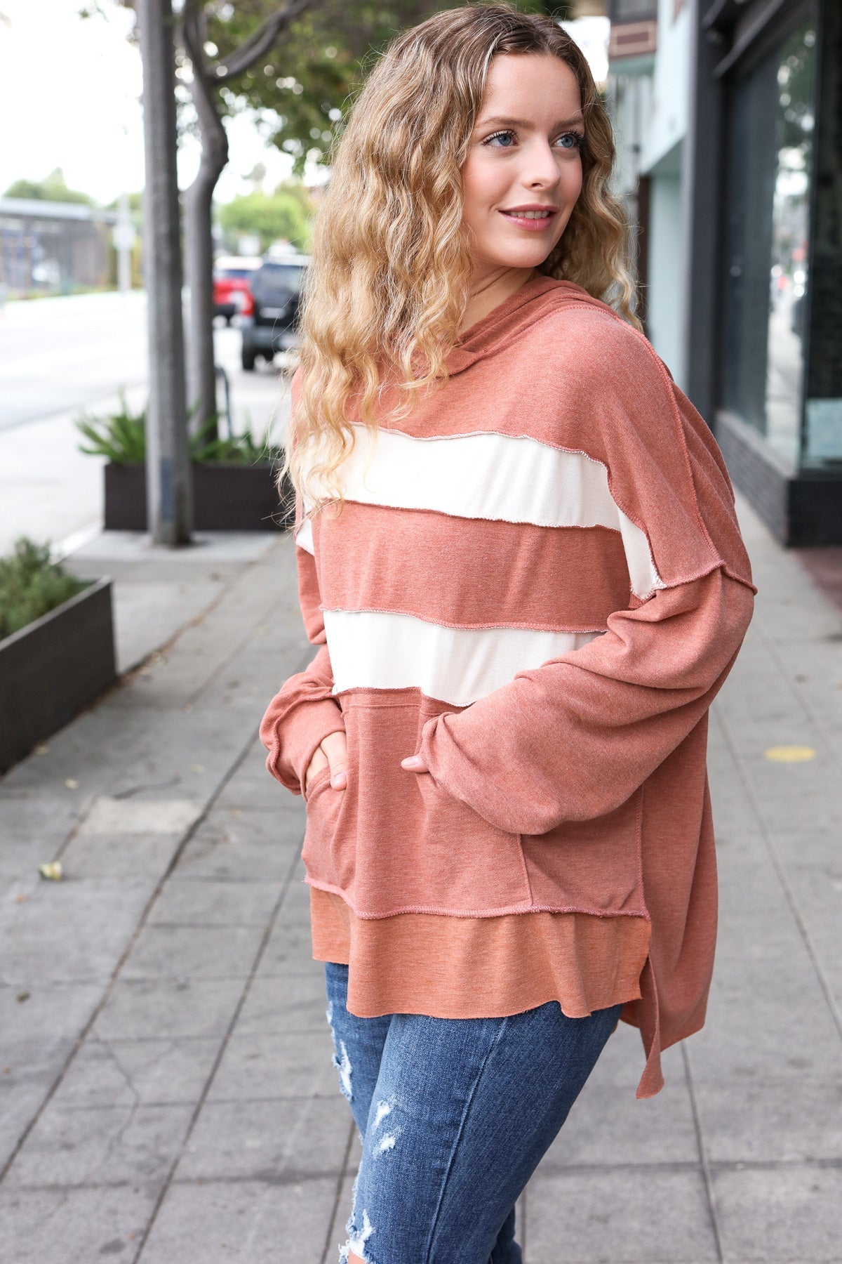 Woman with loose wavy hair wearing a rust-colored hoodie with wide white stripes and distressed jeans outdoors on sidewalk