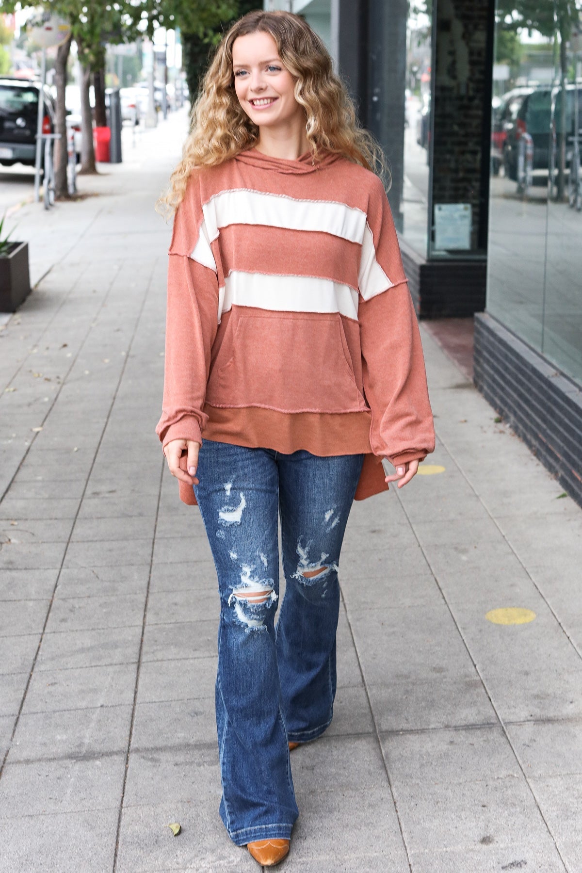 Smiling woman with wavy hair wearing a rust-colored hoodie with wide cream horizontal stripes and distressed blue jeans outdoors