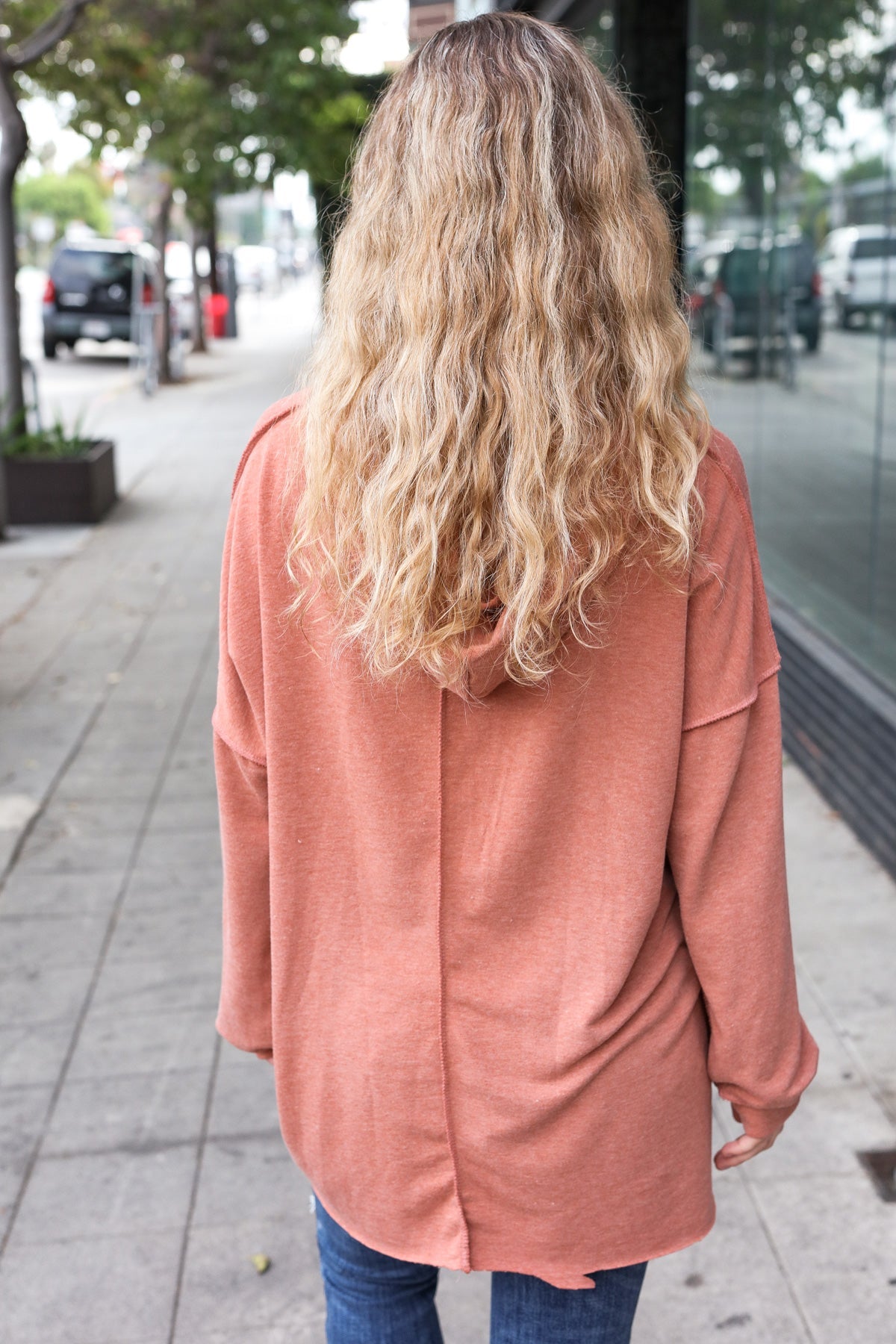 Woman with long wavy blonde hair wearing a loose rust-colored hoodie standing on an urban sidewalk outdoors