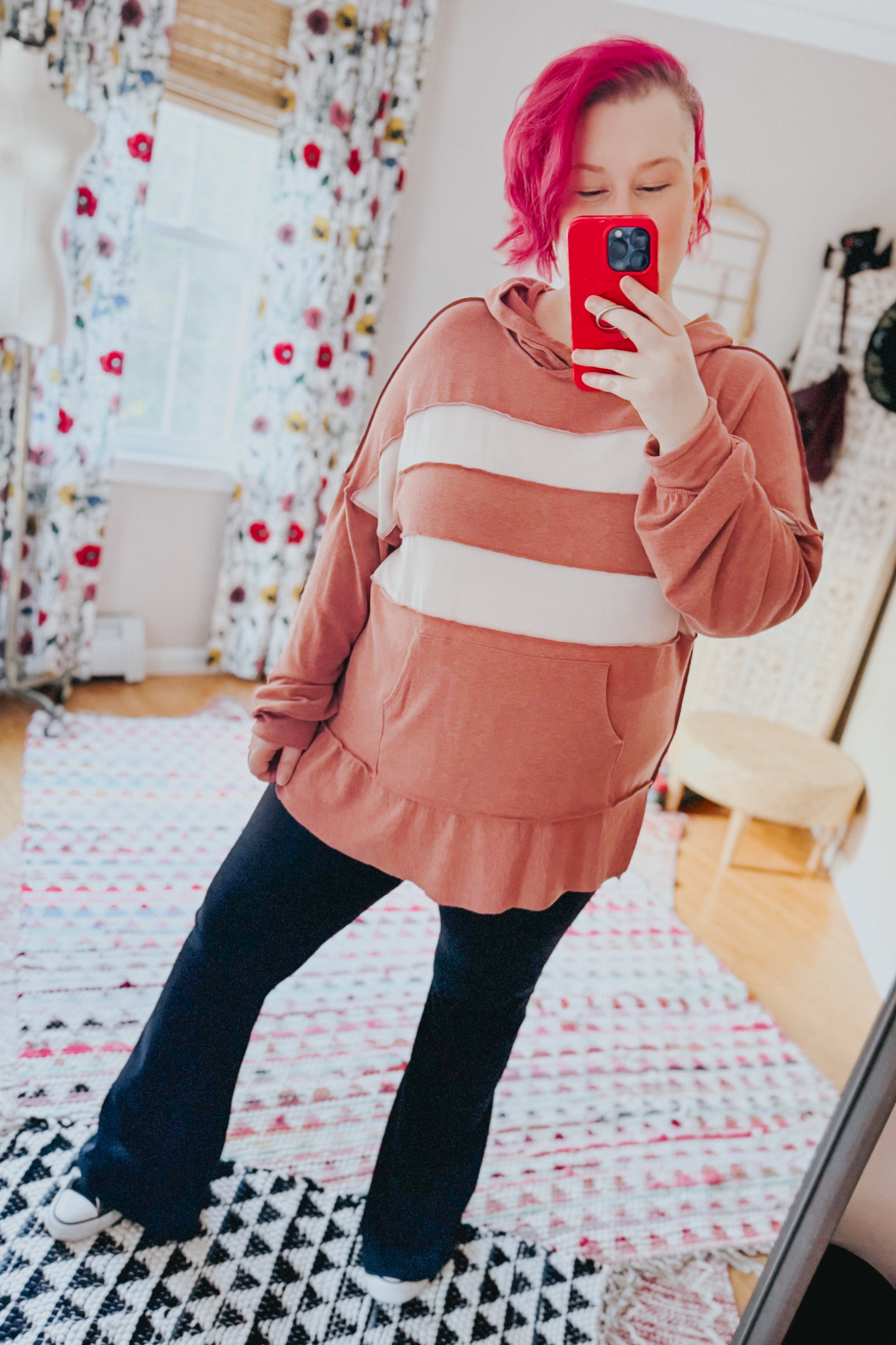 Woman with short pink hair wearing a rust-colored hoodie with two wide cream horizontal stripes and black pants taking a mirror selfie indoors